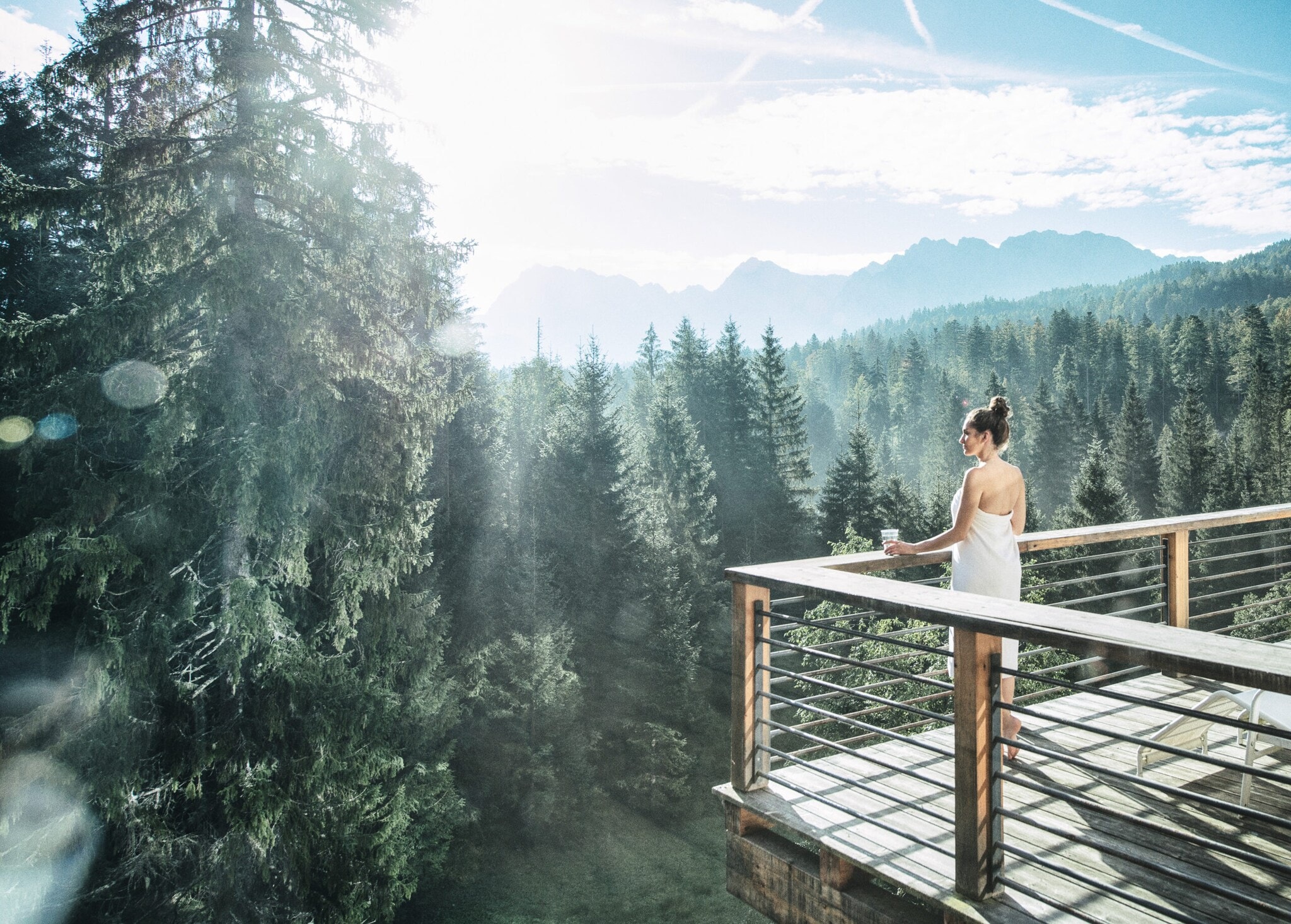 Eine in ein Handtuch gewickelte Frau blickt von einer Terrasse auf einen herrlichen Nadelwald Eine in ein Handtuch gewickelte Frau blickt von einer Terrasse auf einen herrlichen Nadelwald