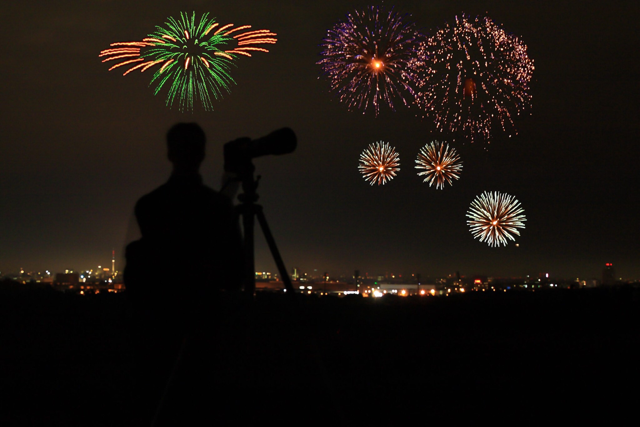 Die Silhouette eines Mannes vor einer Kamera vor einem Feuerwerk über einer Stadt Die Silhouette eines Mannes vor einer Kamera vor einem Feuerwerk über einer Stadt