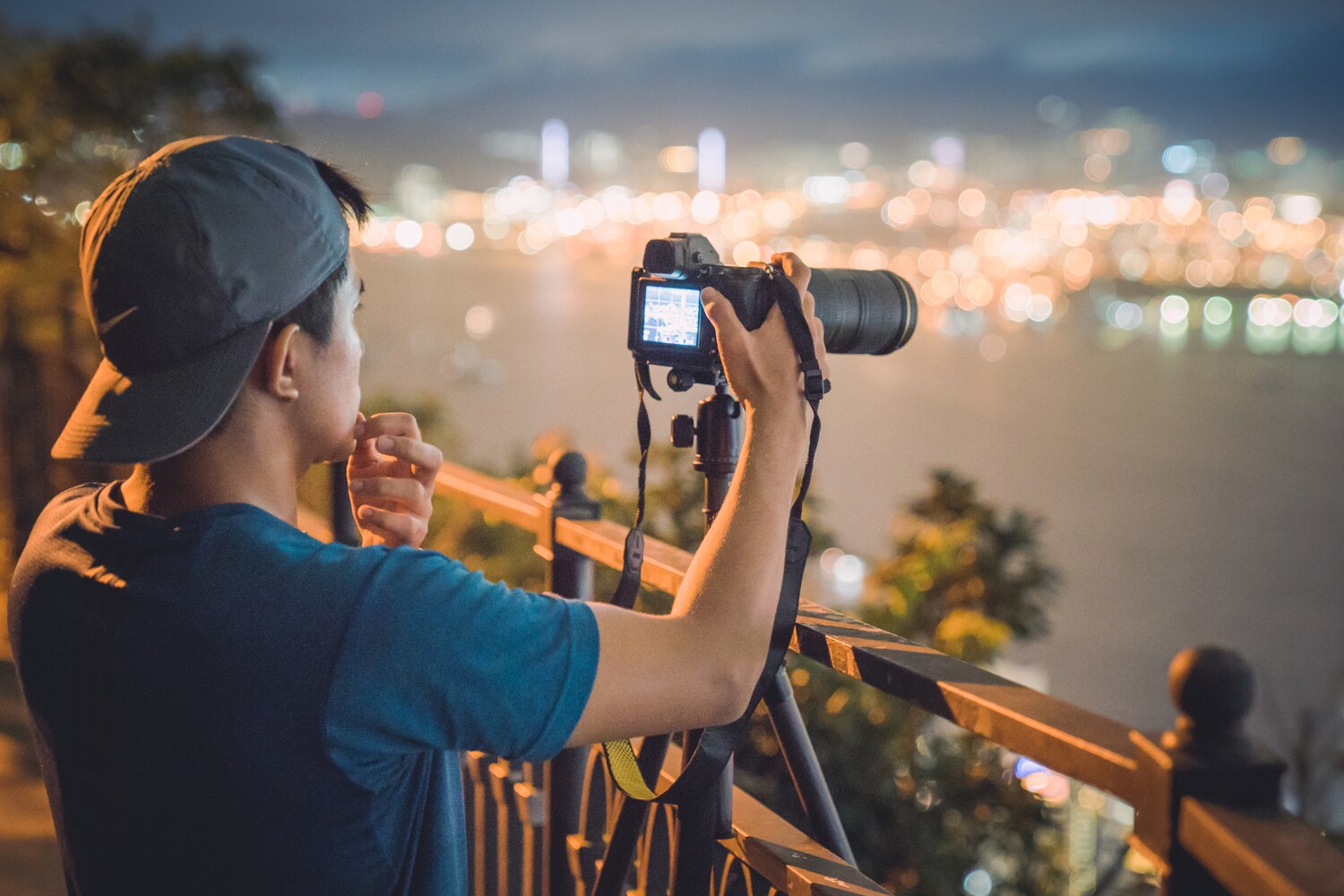 Ein Mann mit Käppi steht vor einem Geländer über einer Stadt und stellt etwas an einem Fotoapparat auf einem Stativ ein Ein Mann mit Käppi steht vor einem Geländer über einer Stadt und stellt etwas an einem Fotoapparat auf einem Stativ ein