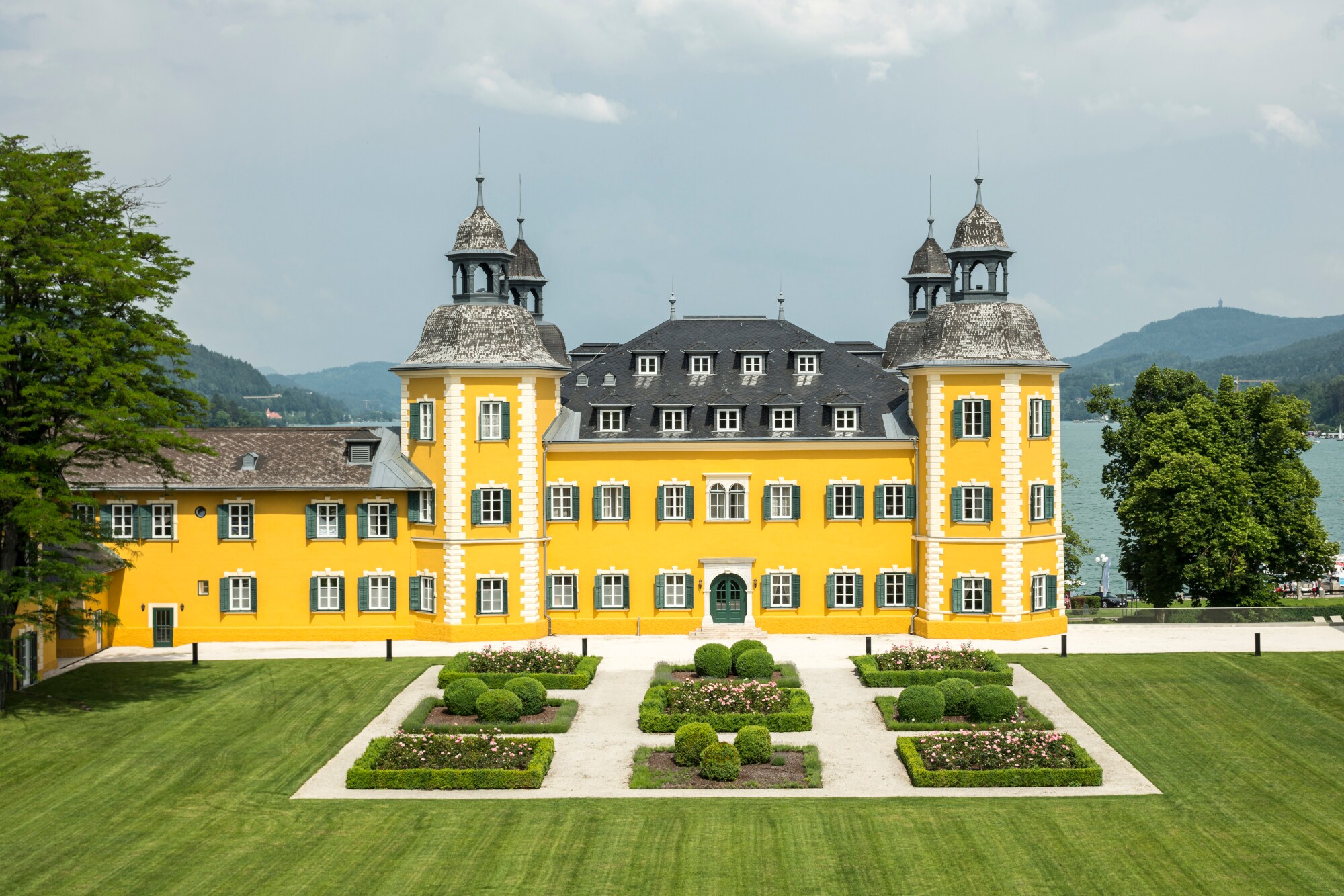 Ein gelbes Schlosshotel vor dem Panorama des Wörthersees