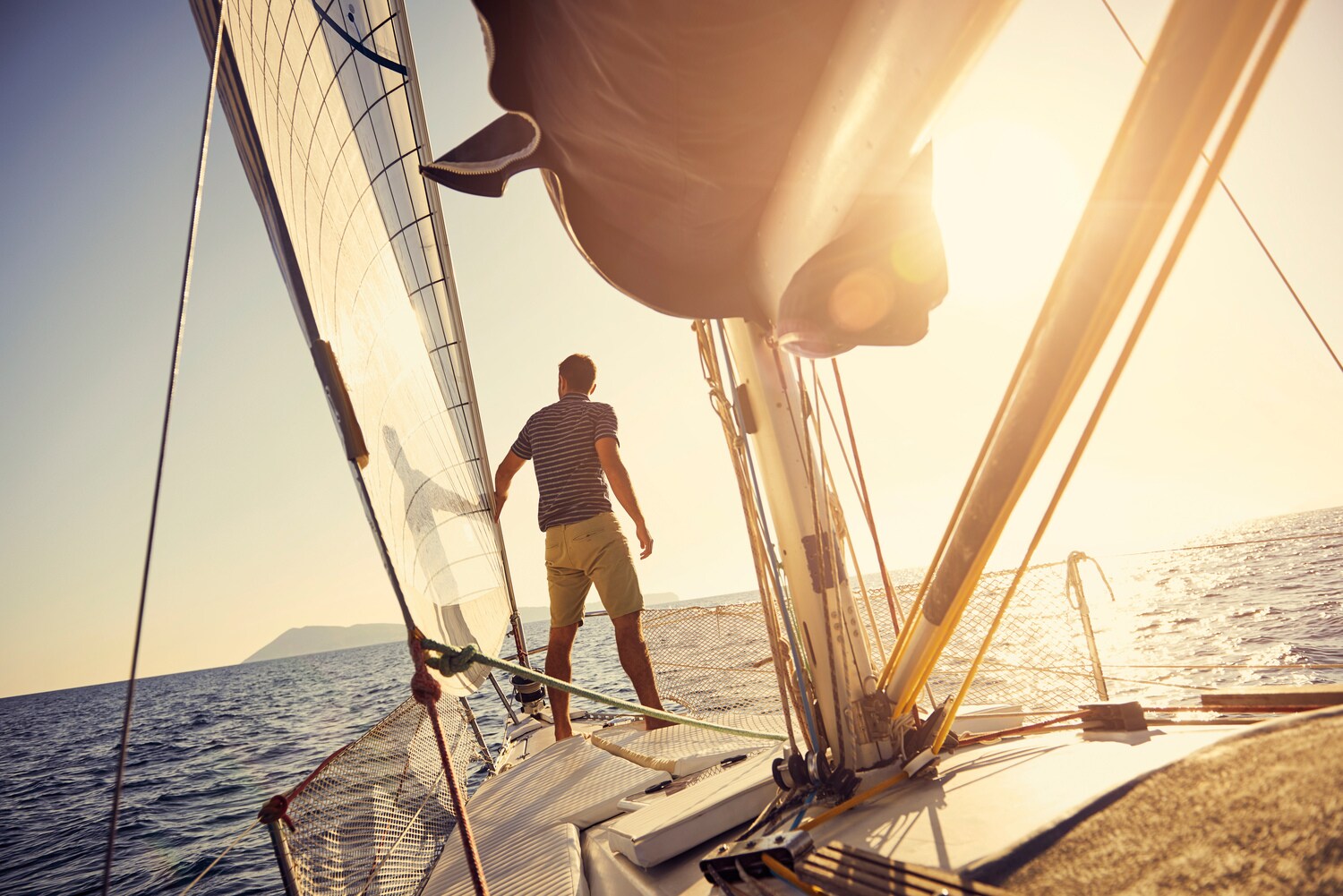 Ein Mann steht am Bug einer Jacht, die bei Sonnenuntergang auf dem Meer in Richtung einer Insel segelt