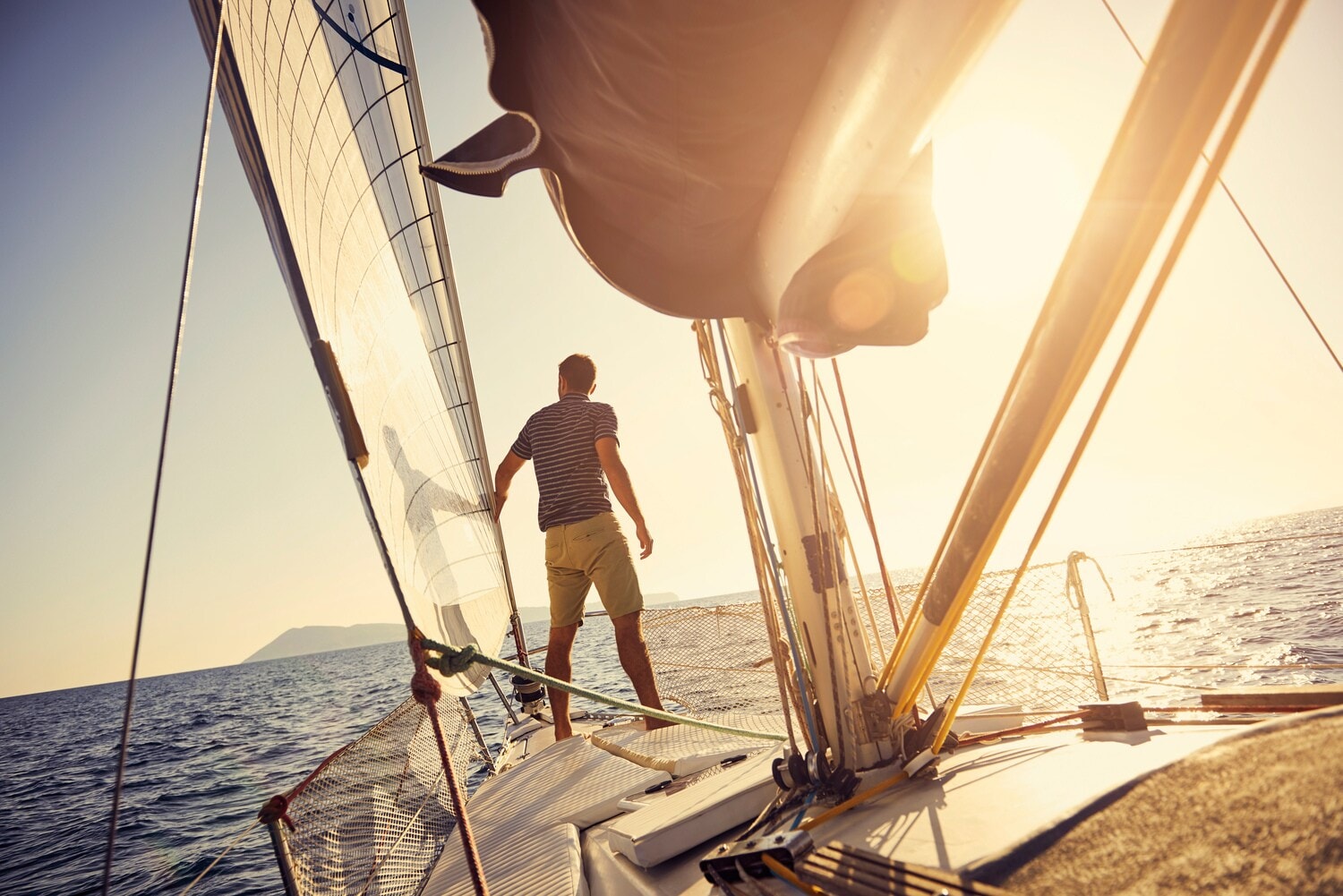 Ein Mann steht am Bug einer Jacht, die bei Sonnenuntergang auf dem Meer in Richtung einer Insel segelt Ein Mann steht am Bug einer Jacht, die bei Sonnenuntergang auf dem Meer in Richtung einer Insel segelt