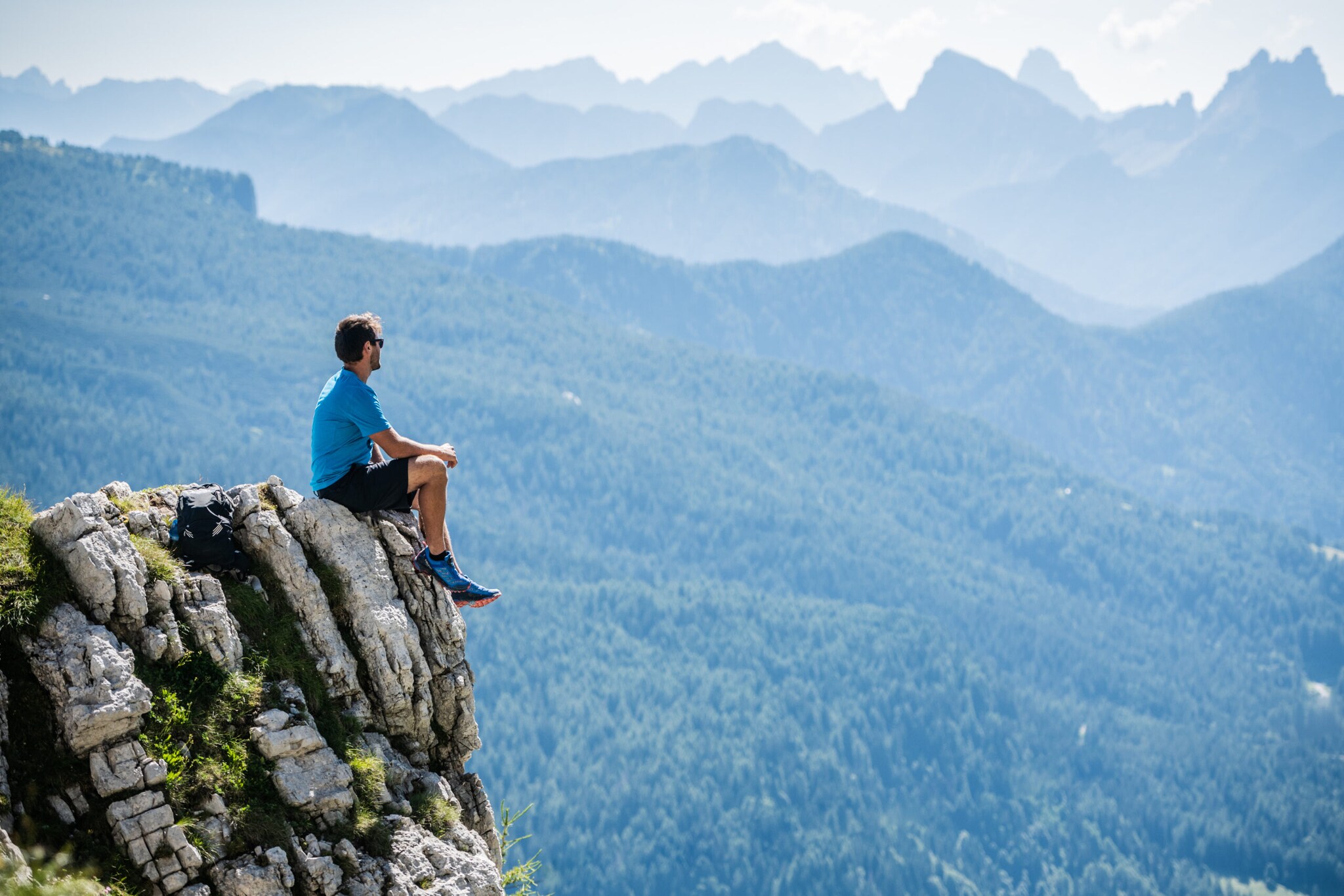 Ein Mann in Sportkleidung sitzt auf Felsvorsprung vor Bergpanorama