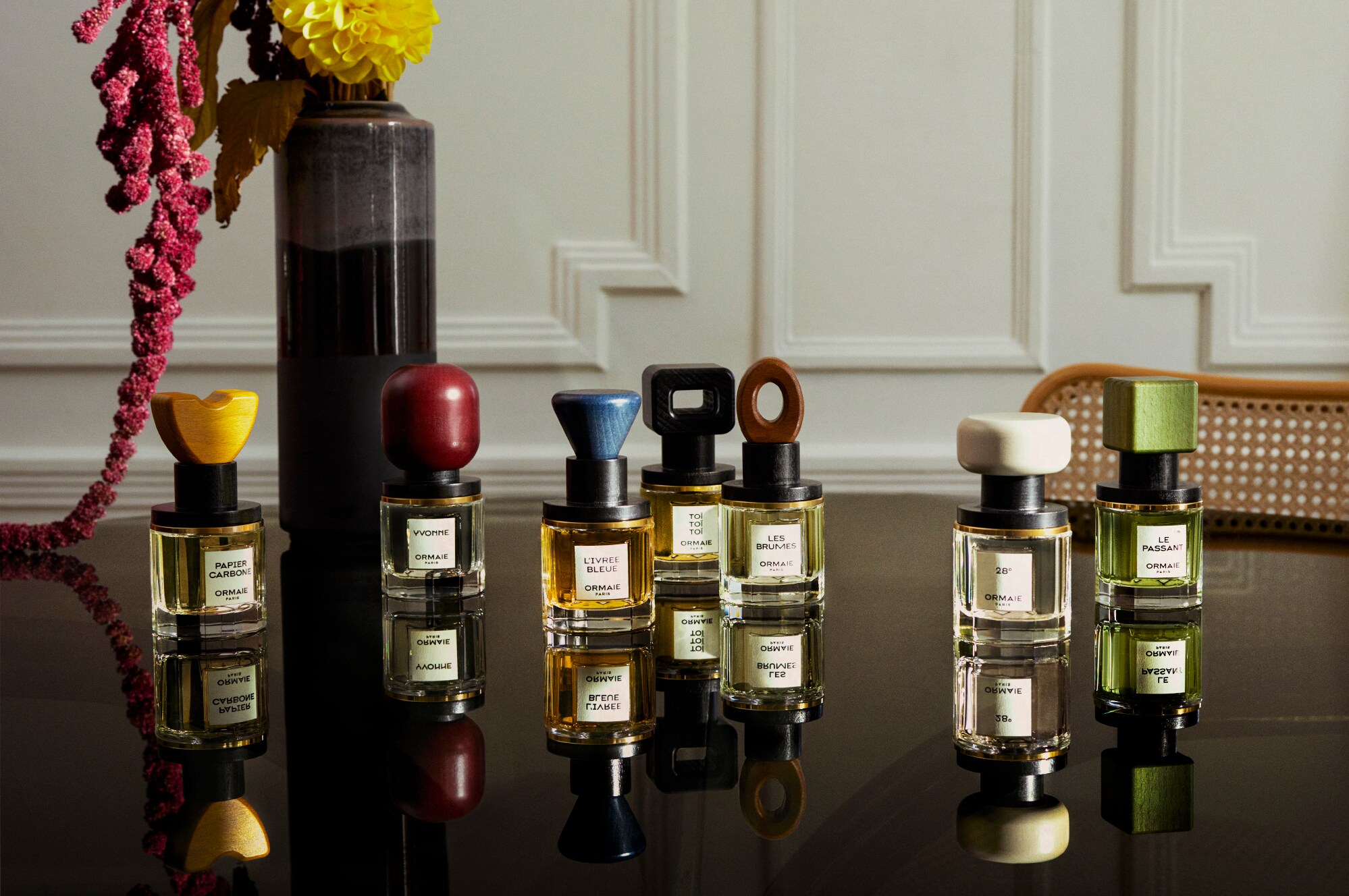 Sieben unterschiedliche Parfümflakons stehen auf einem edlen Tisch mit Blumenvase vor einer weißen Holzvertäfelung