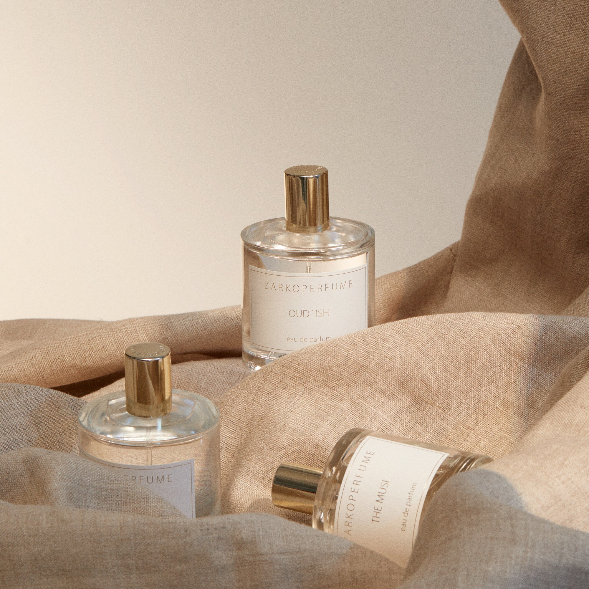 Drei schlichte Parfümflakons mit goldener Kappe sind auf einem beigefarbenem Leinentuch drapiert