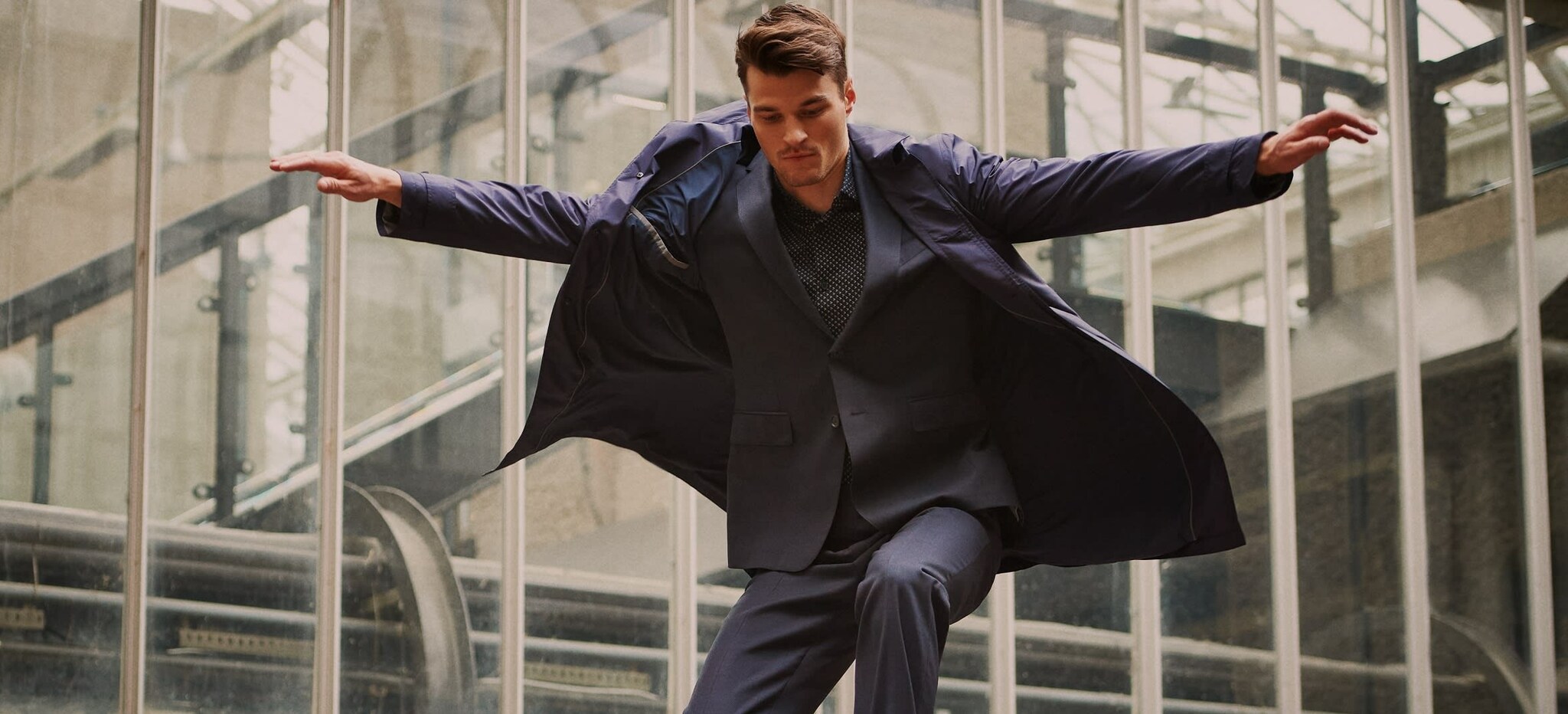 Ein Mann in dunklem Anzug, Mantel und Sneakern bei der Landung eines Sprungs.