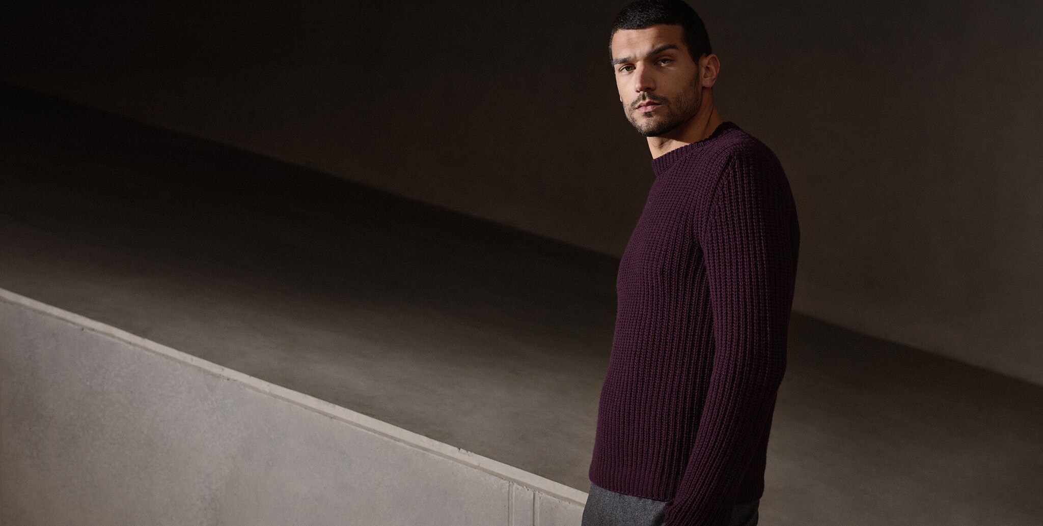 Ein männliches Model in Stoffhose und auberginefarbenem Strickpulli vor einem greigefarbenen Betonhintergrund