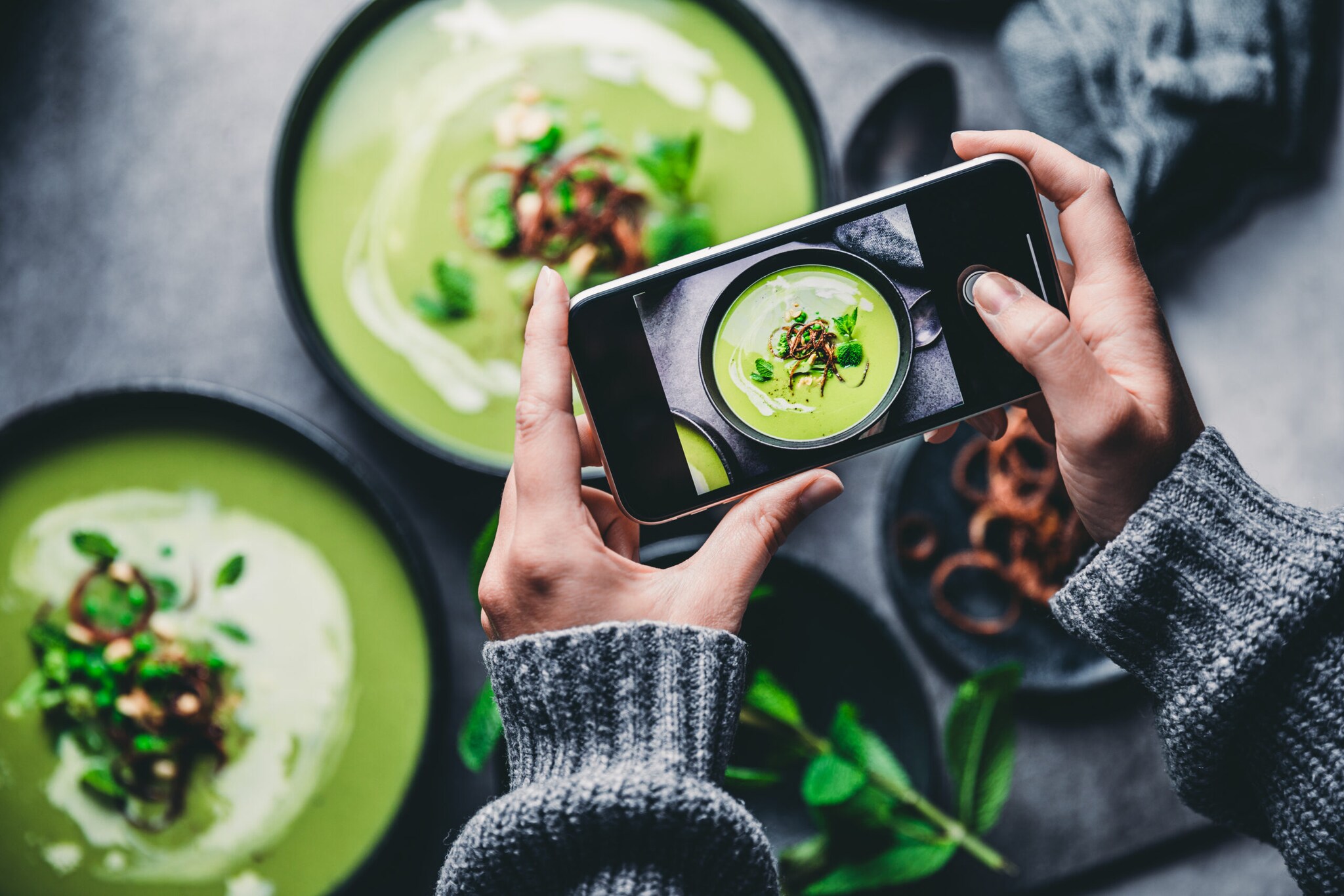 Zwei Hände halten Smartphone, das eine grüne Suppe in Schüssel fotografiert Zwei Hände halten Smartphone, das eine grüne Suppe in Schüssel fotografiert