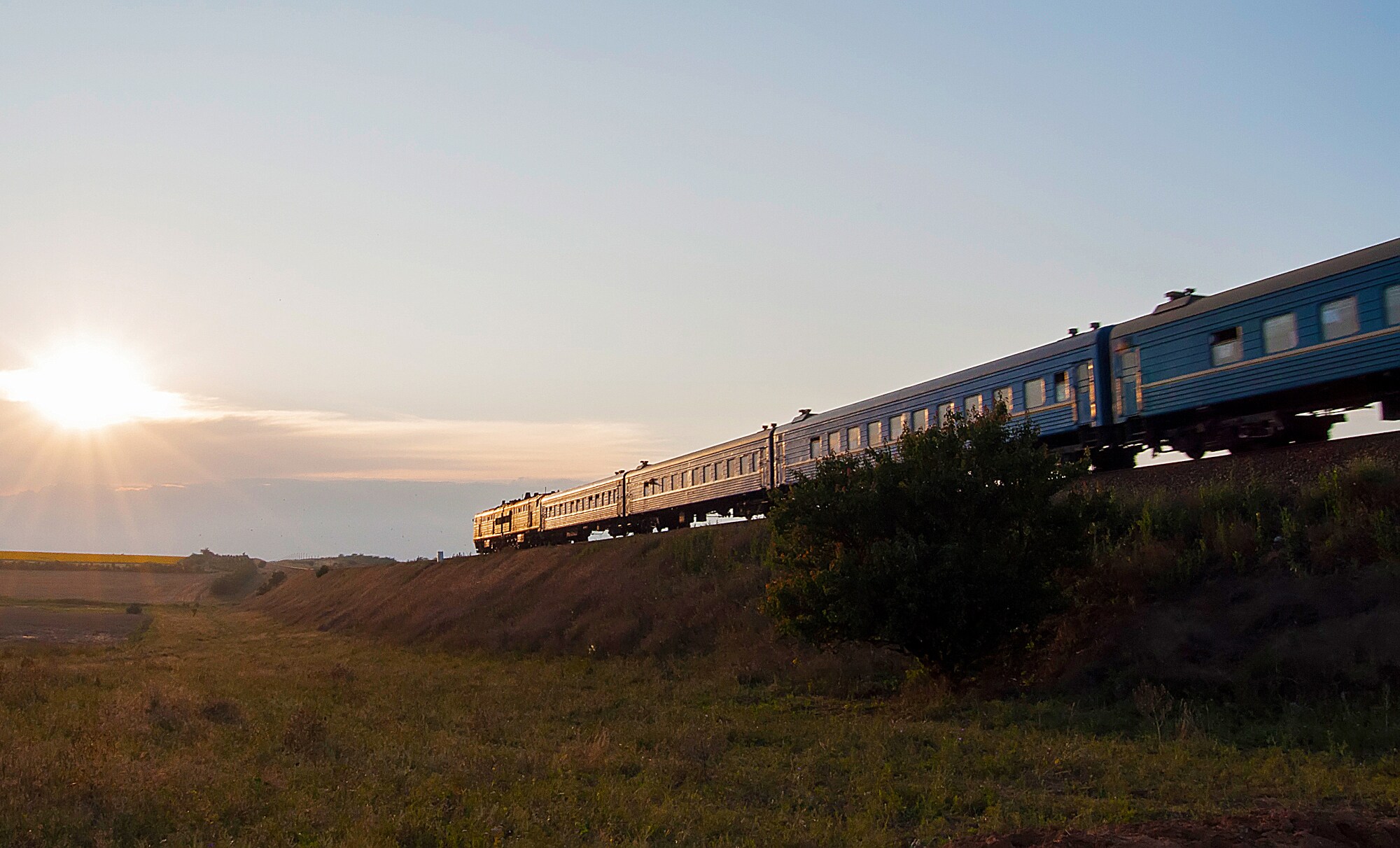 Ein blauer Zug fährt durch Feldlandschaft der Sonne entgegen