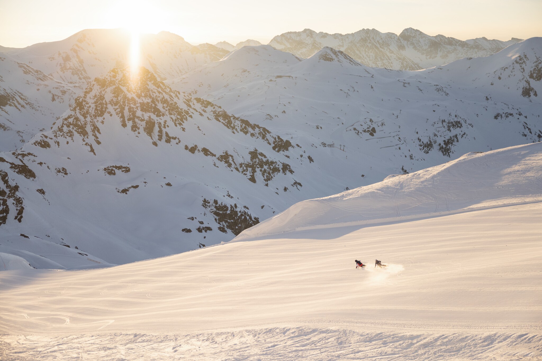 Zwei Skifahrer:innen fahren eine Piste hinab, im Hintergrund eine Bergkette