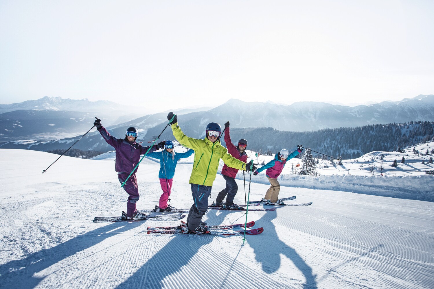 Fünf Skifahrende in bunter Kleidung winken in die Kamera auf einer Piste vor winterlichem Bergpanorama