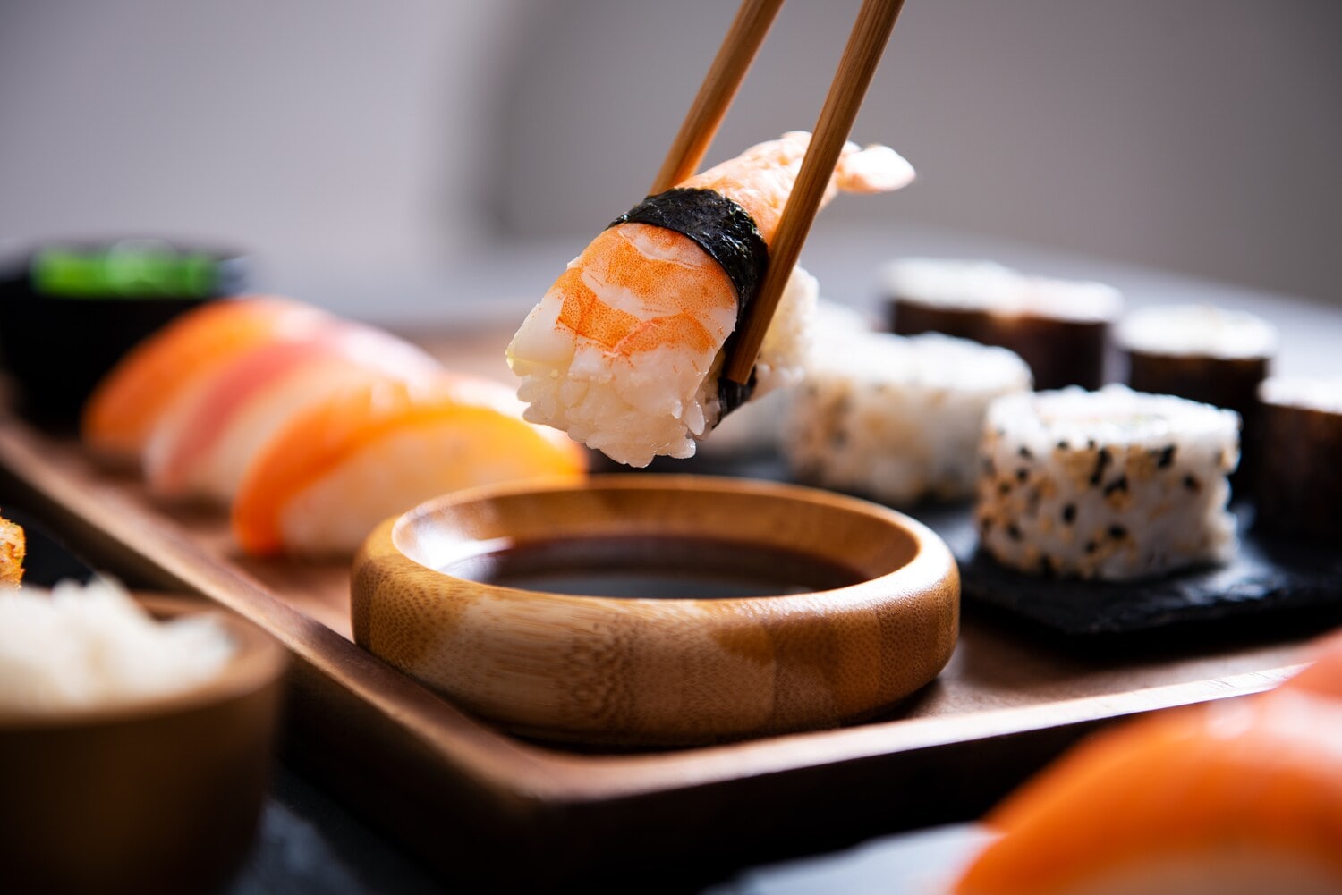 Ein Stück Nigiri-Sushi mit Garnele wird von zwei Essstäbchen über einer Holzschale mit Sojasauce gehalten, im Hintergrund weitere Sushi-Stücke auf Tablett Ein Stück Nigiri-Sushi mit Garnele wird von zwei Essstäbchen über einer Holzschale mit Sojasauce gehalten, im Hintergrund weitere Sushi-Stücke auf Tablett