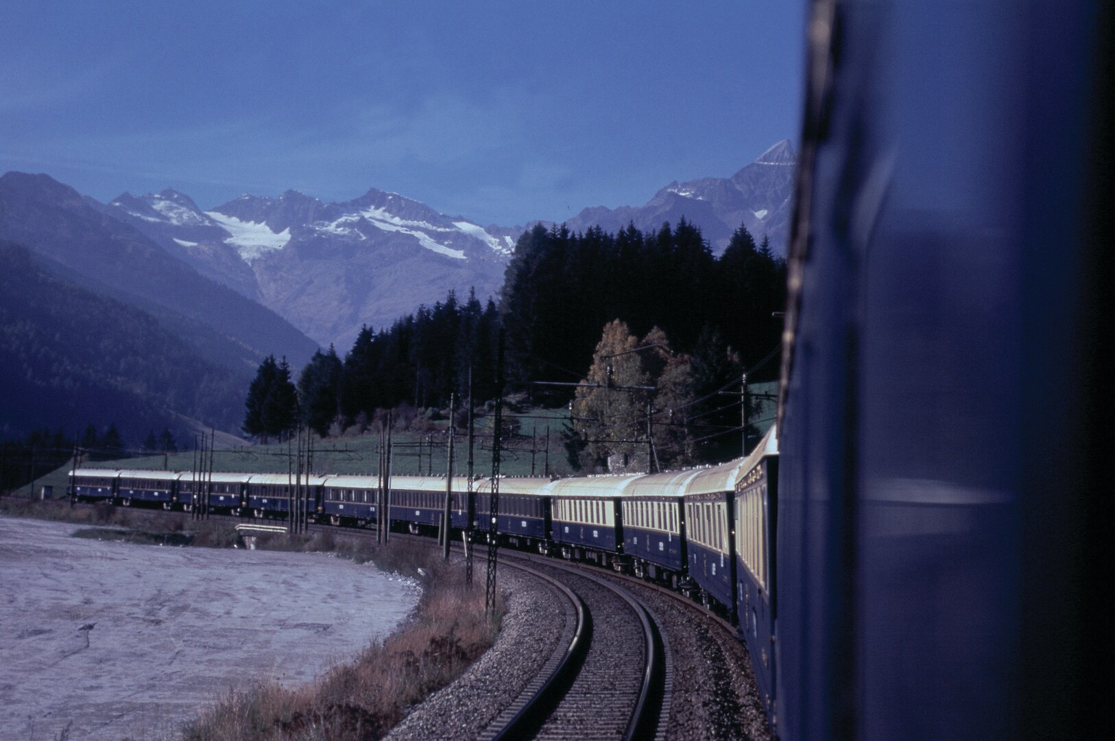 Ein blauer Zug fährt in einer Kurve in der Dämmerung durch die Landschaft, im Hintergrund Wald und Bergpanorama Ein blauer Zug fährt in einer Kurve in der Dämmerung durch die Landschaft, im Hintergrund Wald und Bergpanorama