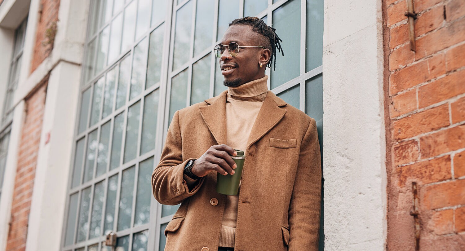 Ein junger, moderner Mann, gekleidet mit beigem Pullover und beigem Wollmantel, lehnt an einem Gebäude mit einem Thermobecher in der Hand