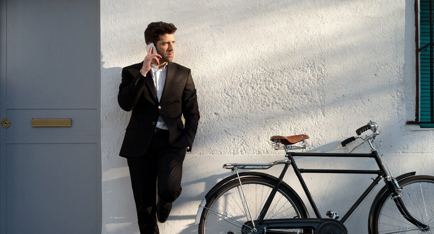 Ein junger Mann im Anzug lehnt telefonierend neben einem Fahrrad an einer Hauswand