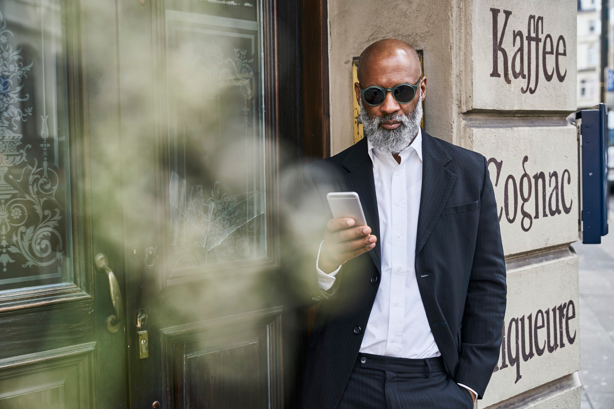 Ein Mann in Anzug und mit einer runden Sonnenbrille hat ein Handy in der Hand und lehnt an einer Hauswand