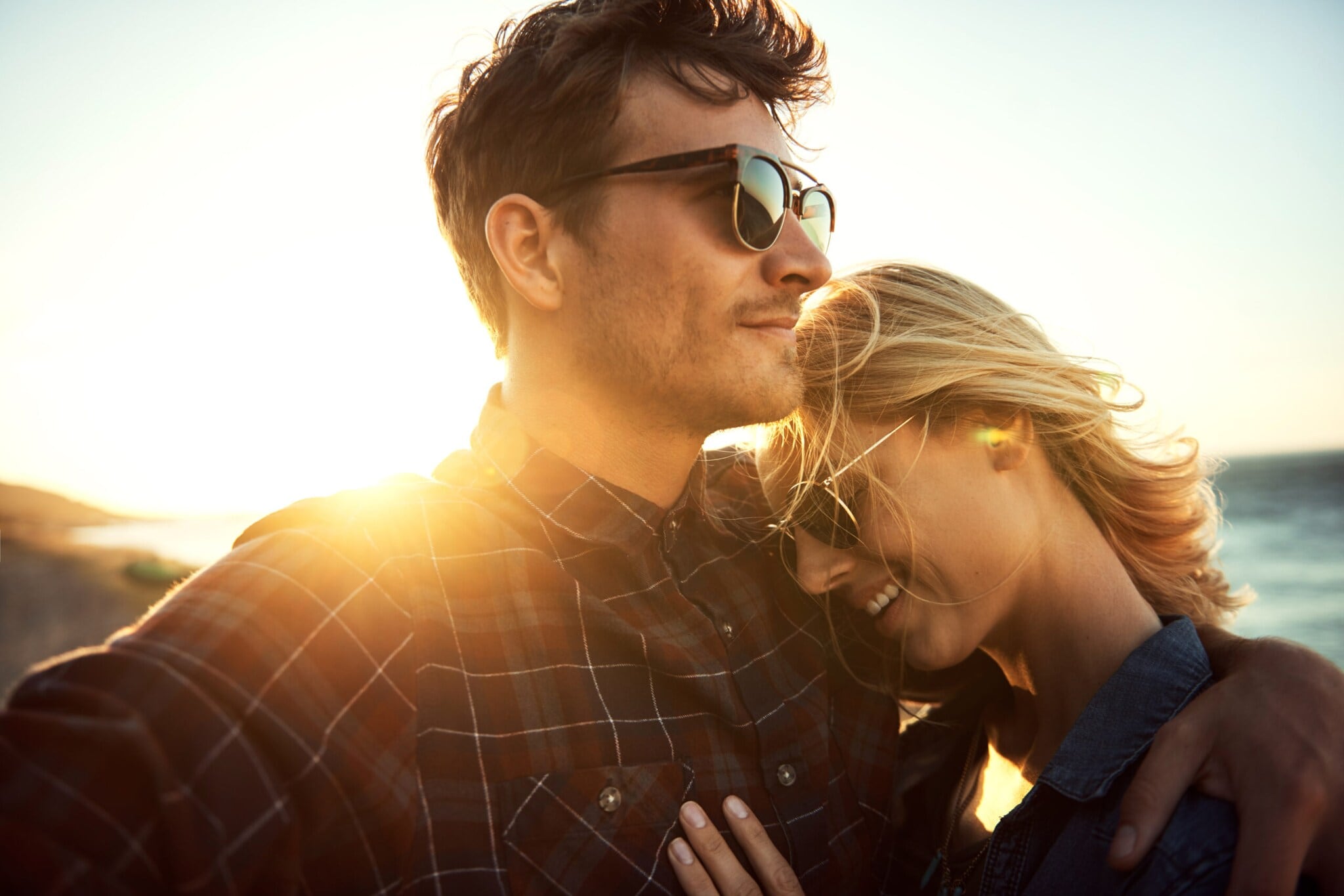 Eine Frau lehnt sich an ihren Partner, sie tragen beide eine Sonnenbrille, im Hintergrund sind das Meer und die untergehende Sonne zu erkennen Eine Frau lehnt sich an ihren Partner, sie tragen beide eine Sonnenbrille, im Hintergrund sind das Meer und die untergehende Sonne zu erkennen