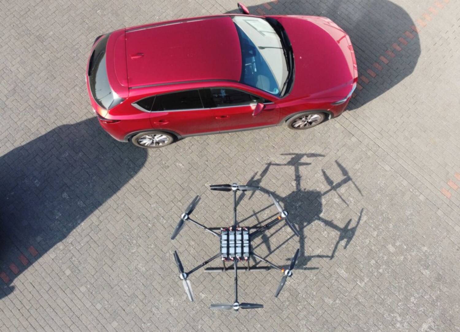 Eine große sechsarmige Drohne steht neben einem roten Kleinwagen und hat in etwa die gleiche Breite