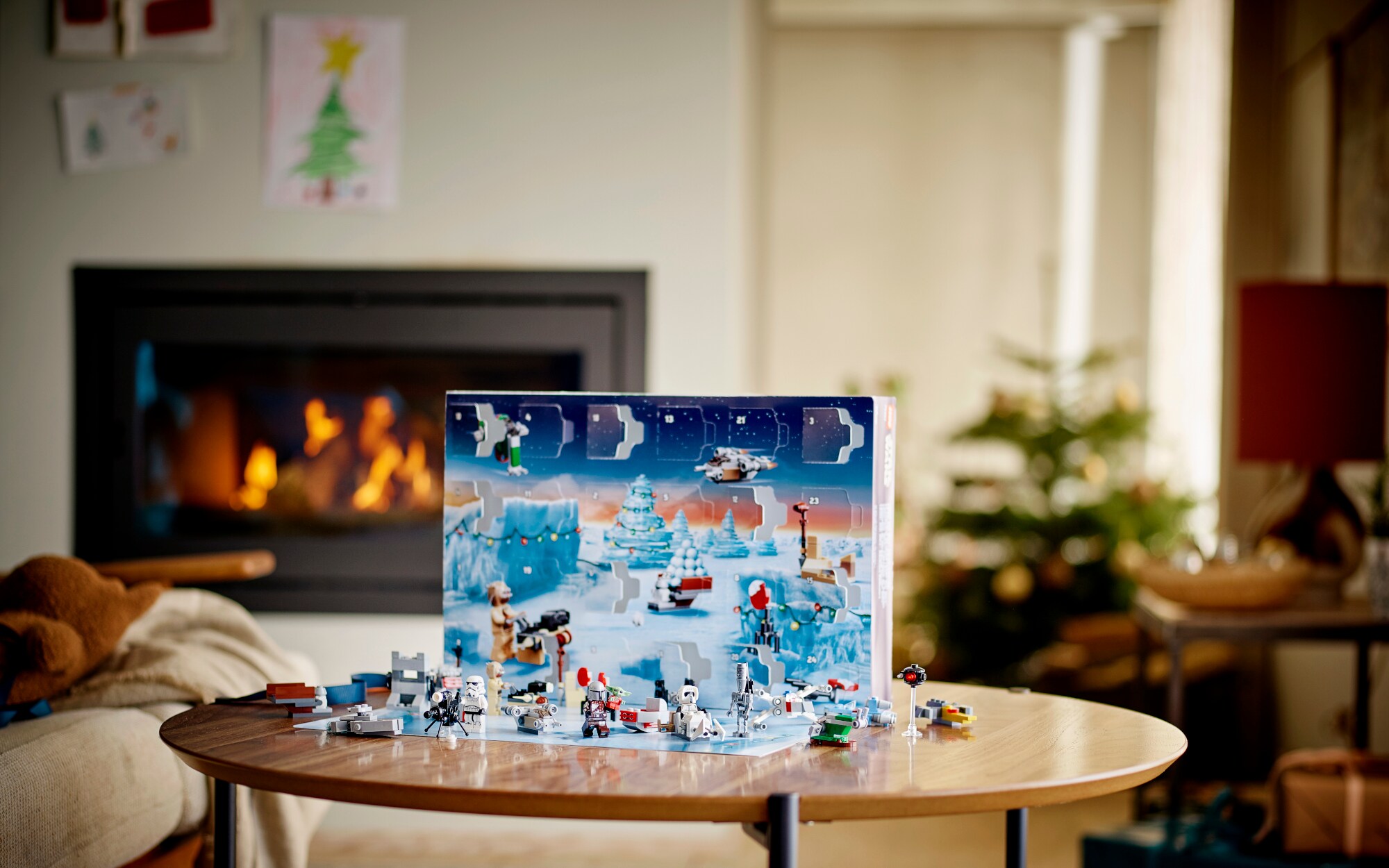 Ein geöffneter Adventskalender mit aufgebauten Lego-Spielfiguren steht auf einem Tisch in einem weihnachtlich geschmückten Wohnzimmer mit Kamin