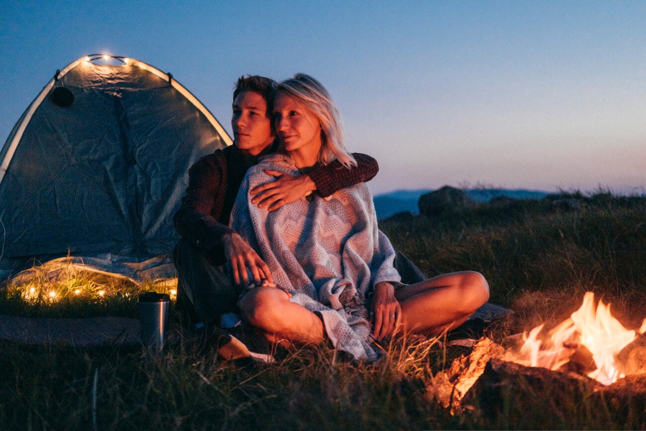 Ein Paar sitzt in der Dämmerung vor einem Zelt, ein Lagerfeuer brennt.