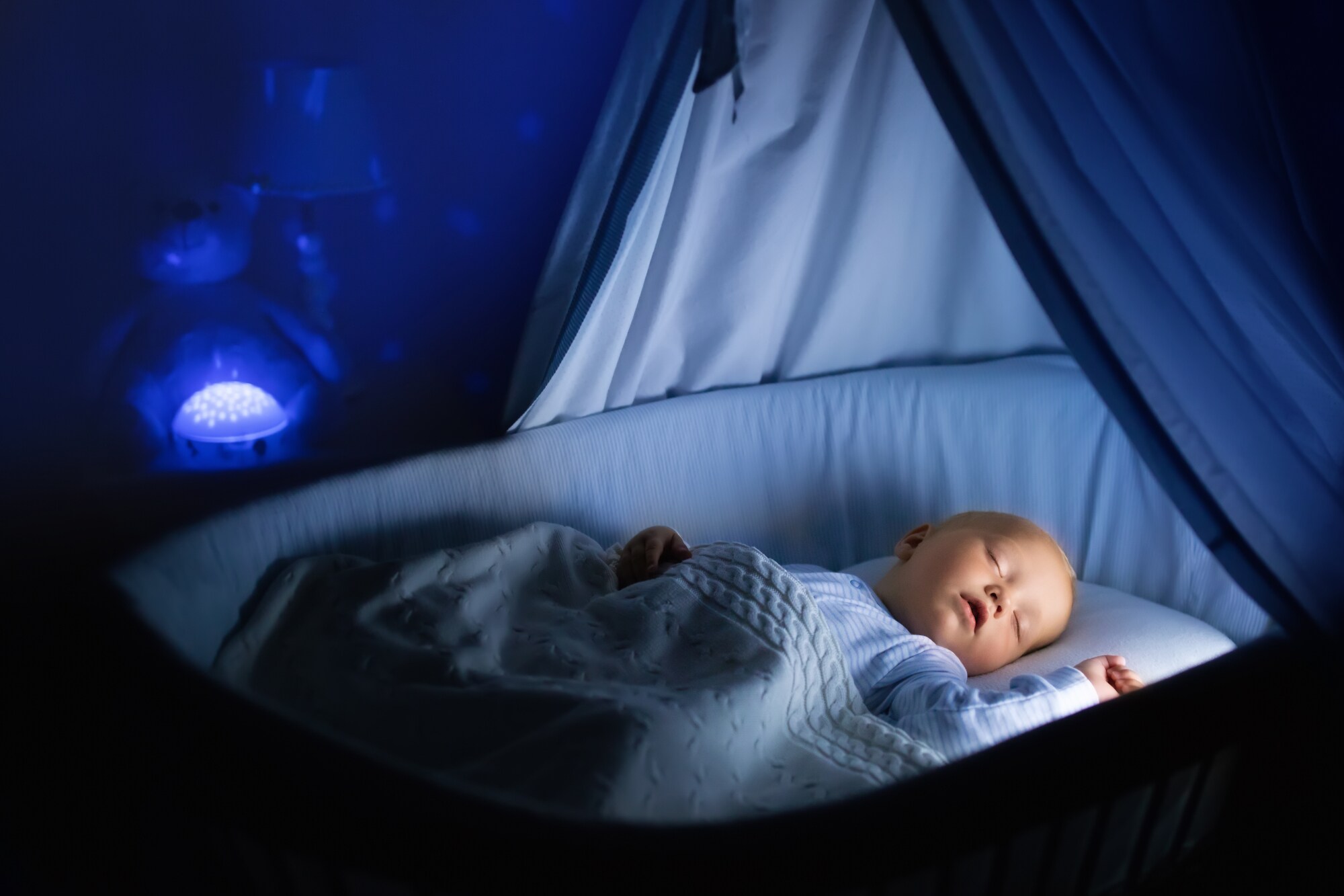 Ein Baby schläft im beleuchteten Babybett in einem dunklen Raum, im Hintergrund blaues Lichtspiel an der Wand
