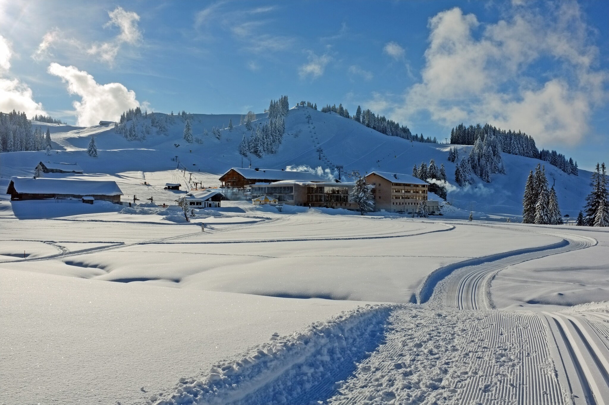 Loipen neben Neuschnee, im Hintergrund Hütten und ein schneebedeckter Berg.