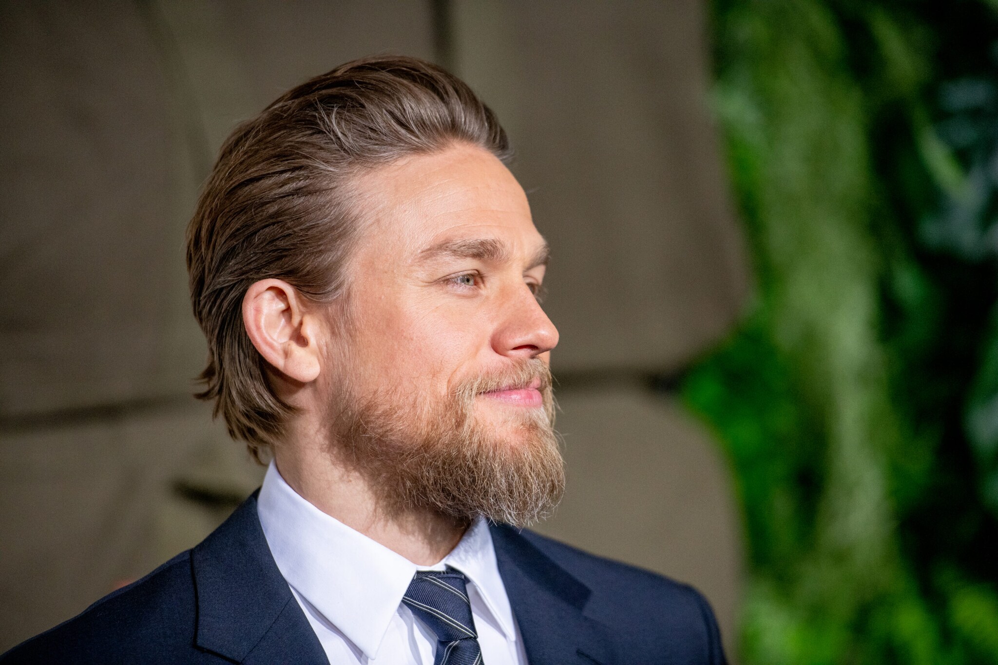 Porträt von Charlie Hunnam mit Vollbart und Anzug vor beige-grünem Hintergrund.
