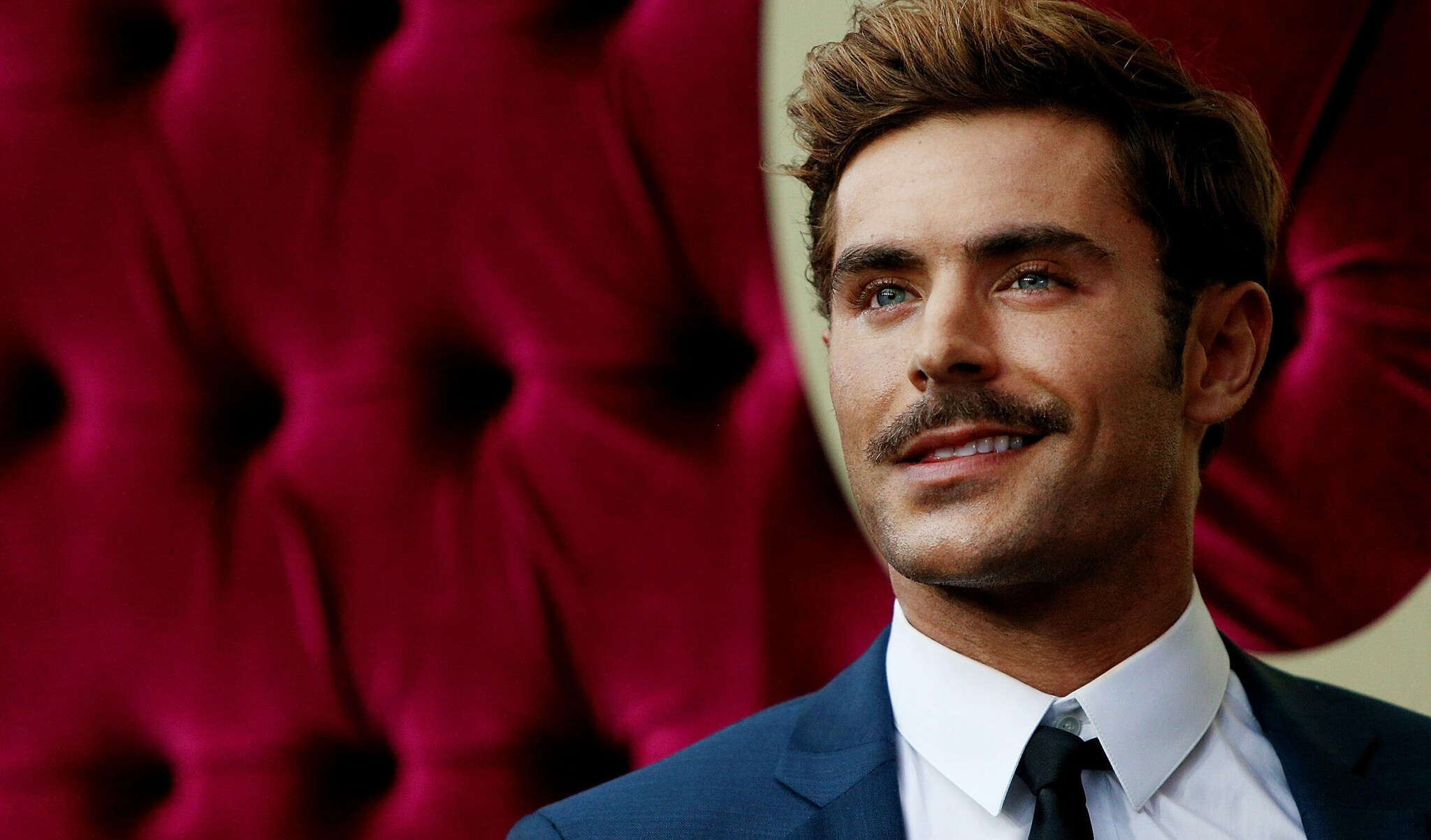 Porträt von Zac Efron mit Schnurrbart und Anzug vor rotem Hintergrund.