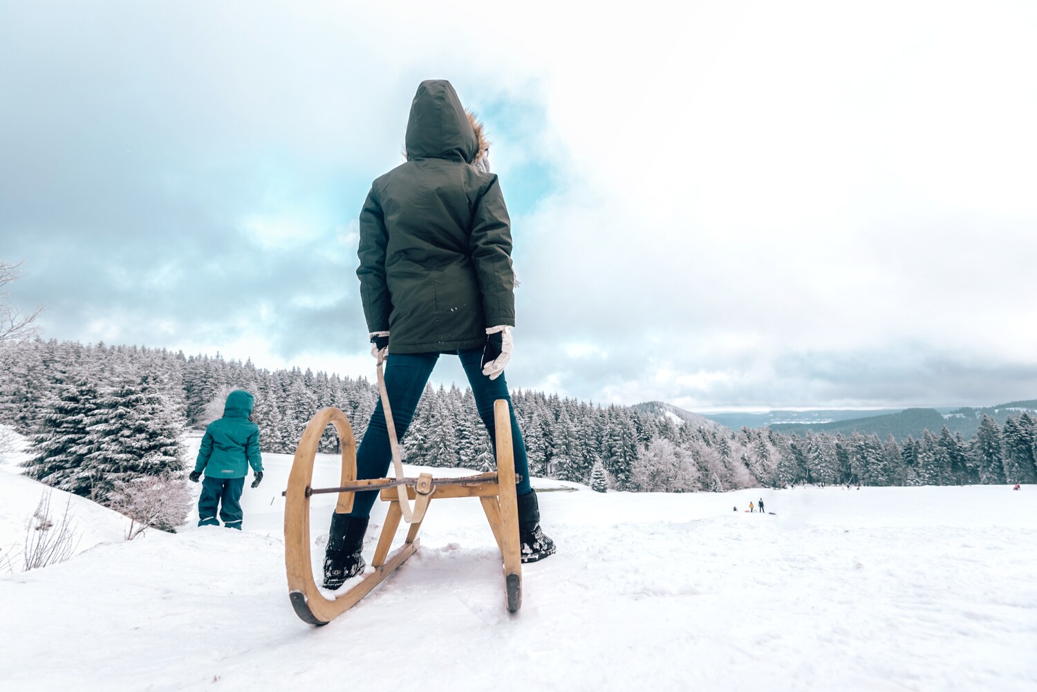 Zwei Kinder mit Schlitten auf einer Schneefläche, im Hintergrund Wälder und Berge.