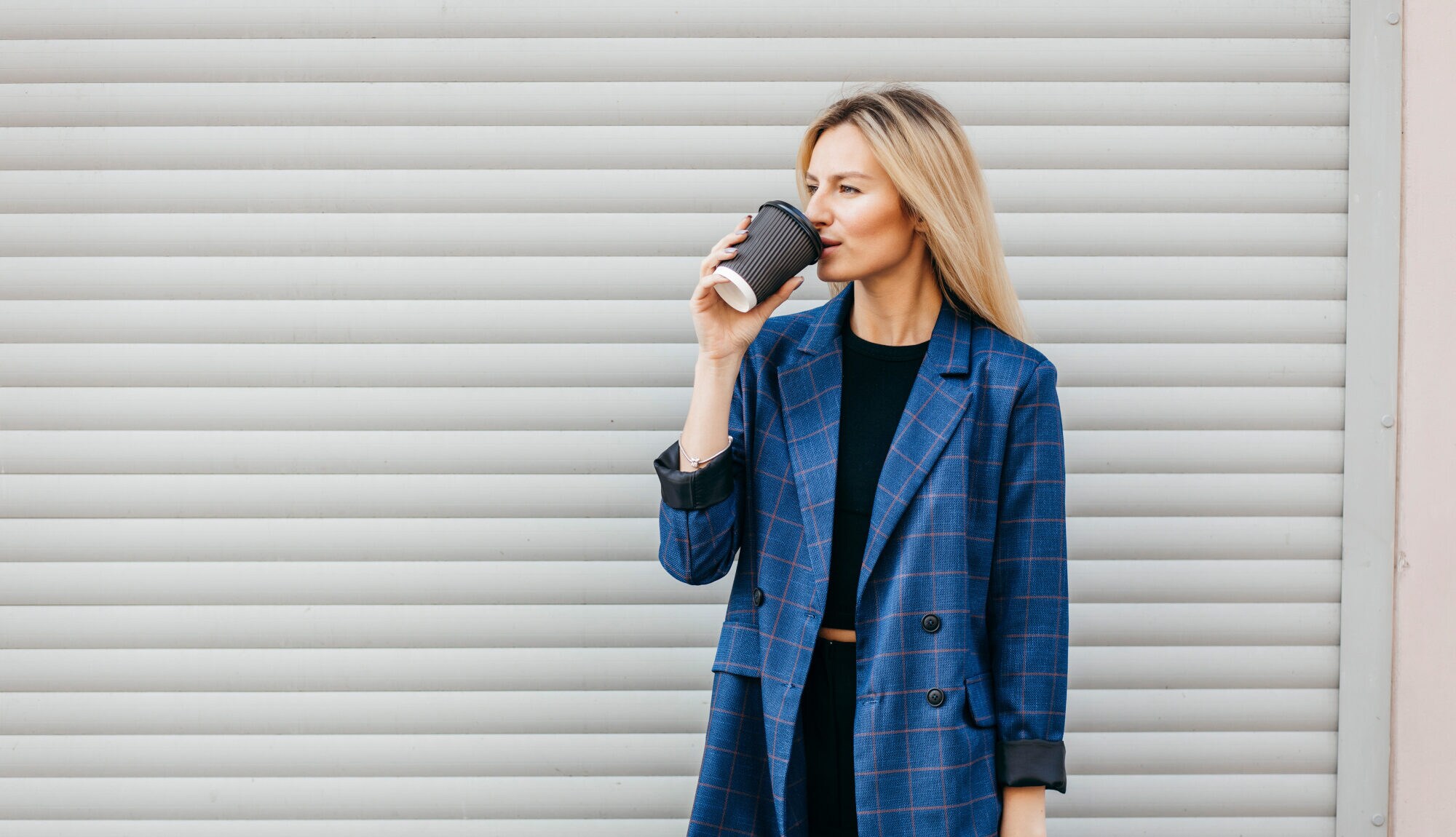 Eine Frau in einem blauen Blazer steht vor einer Garage und trinkt aus einem Becher Eine Frau in einem blauen Blazer steht vor einer Garage und trinkt aus einem Becher