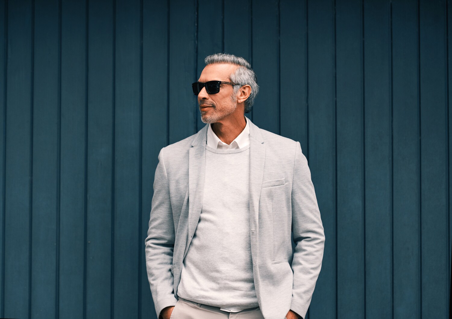 Mann vor einer blauen Holzwand mit weißem Pullover und hellem Anzug mit Sonnenbrille Mann vor einer blauen Holzwand mit weißem Pullover und hellem Anzug mit Sonnenbrille