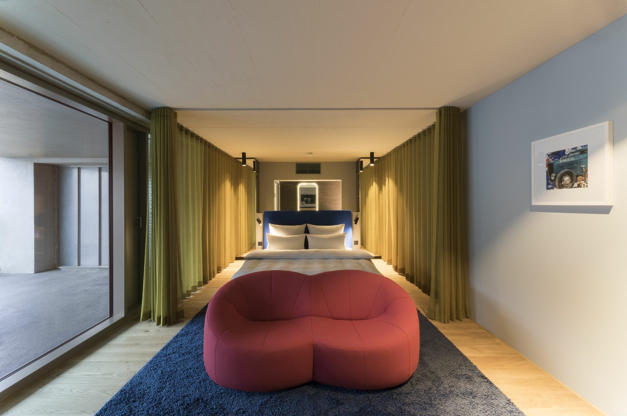 Ein schick eingerichtetes und beleuchtetes Hotelzimmer hat, getrennt durch eine Glaswand, nebenan einen Parkplatz mit Autolift. Ein schick eingerichtetes und beleuchtetes Hotelzimmer hat, getrennt durch eine Glaswand, nebenan einen Parkplatz mit Autolift.