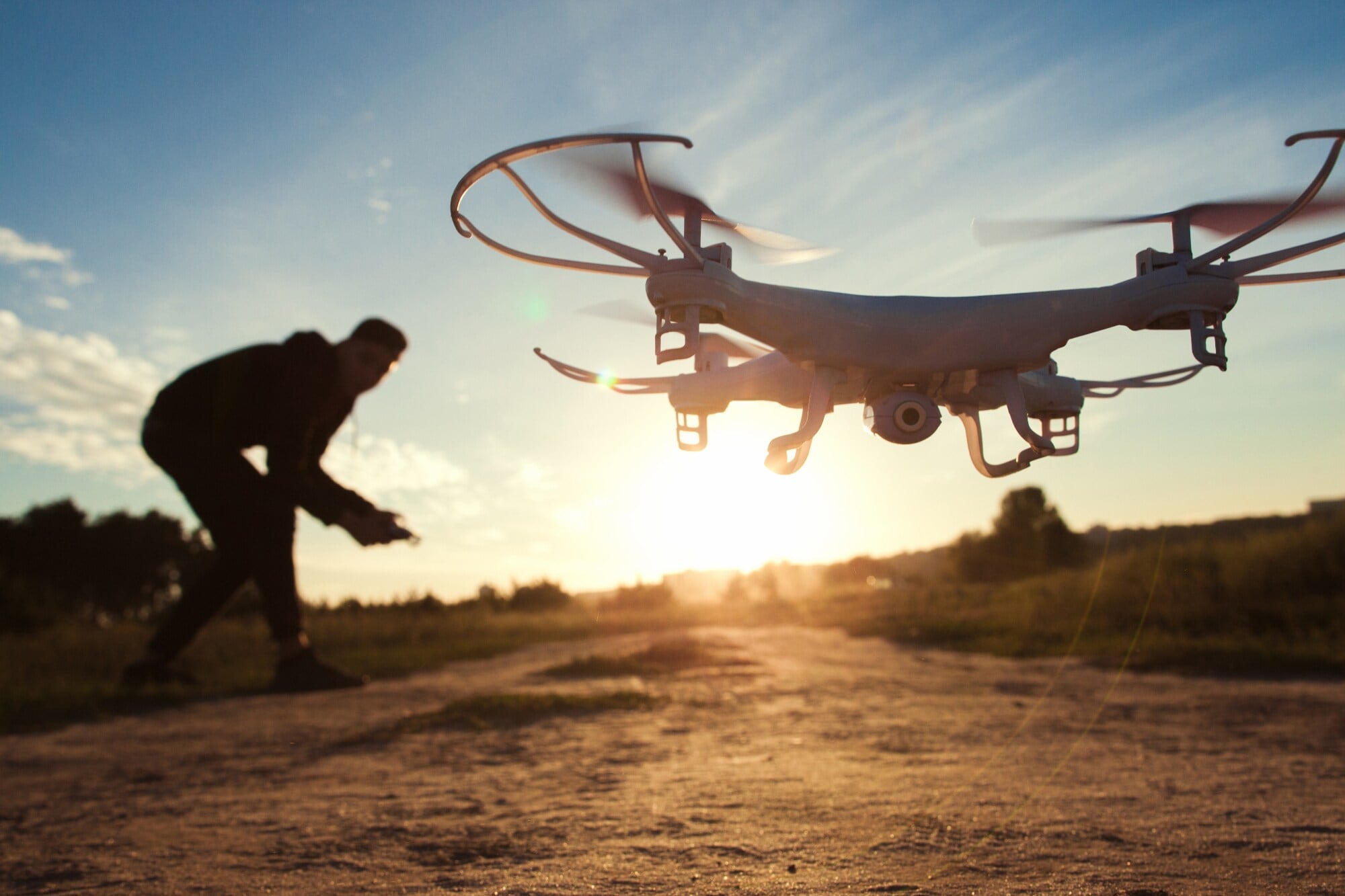 Eine Drohne mit Kamera an der Unterseite fliegt im Tiefflug über ein Feld im Sonnenuntergang, im Hintergrund der Pilot als Silhouette Eine Drohne mit Kamera an der Unterseite fliegt im Tiefflug über ein Feld im Sonnenuntergang, im Hintergrund der Pilot als Silhouette