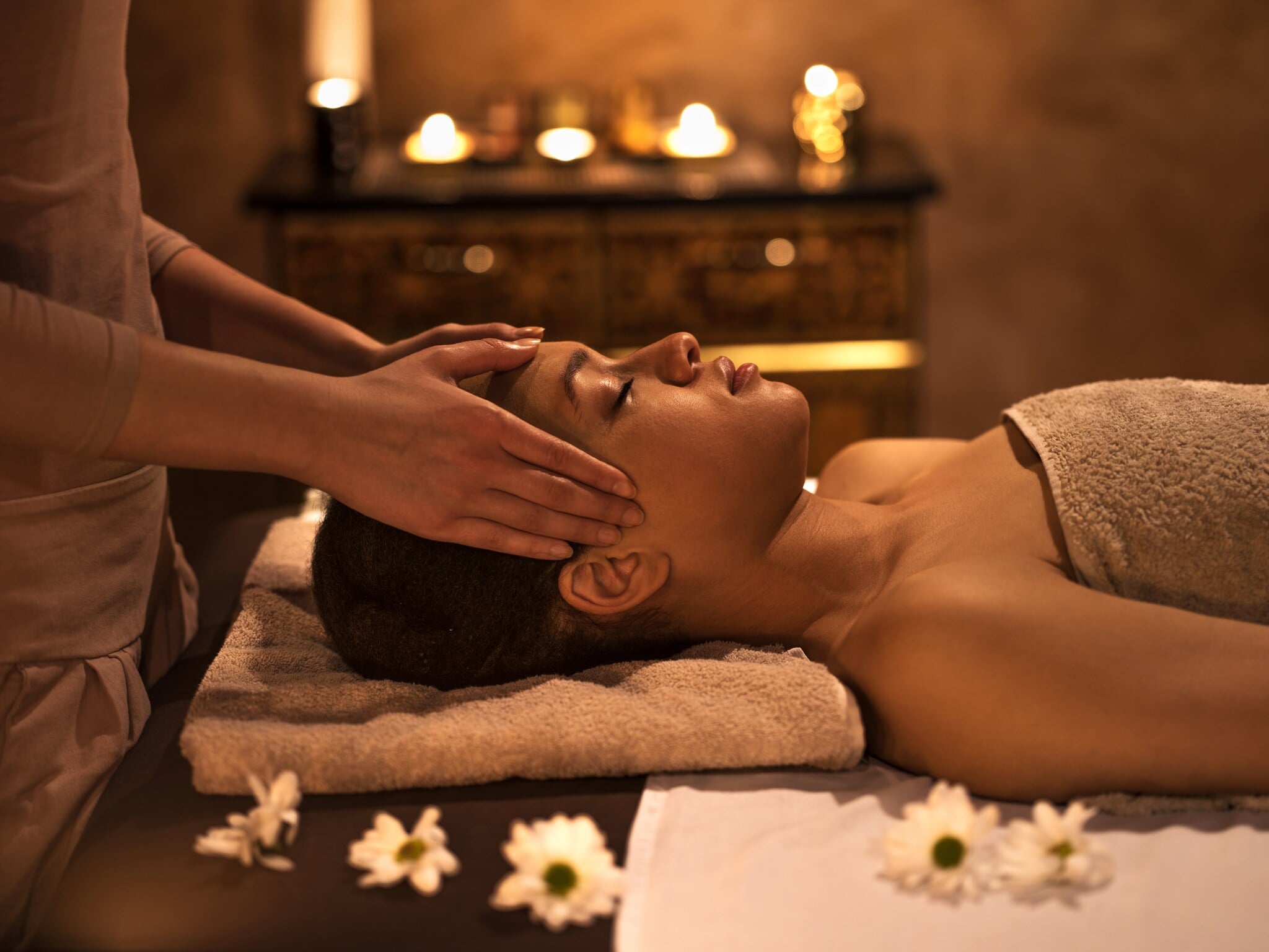 Zwei Hände massieren den Kopf einer jungen Frau, die mit geschlossenen Augen im Handtuch auf einer Liege liegt, drumherum sind Blüten und Kerzen dekoriert Zwei Hände massieren den Kopf einer jungen Frau, die mit geschlossenen Augen im Handtuch auf einer Liege liegt, drumherum sind Blüten und Kerzen dekoriert