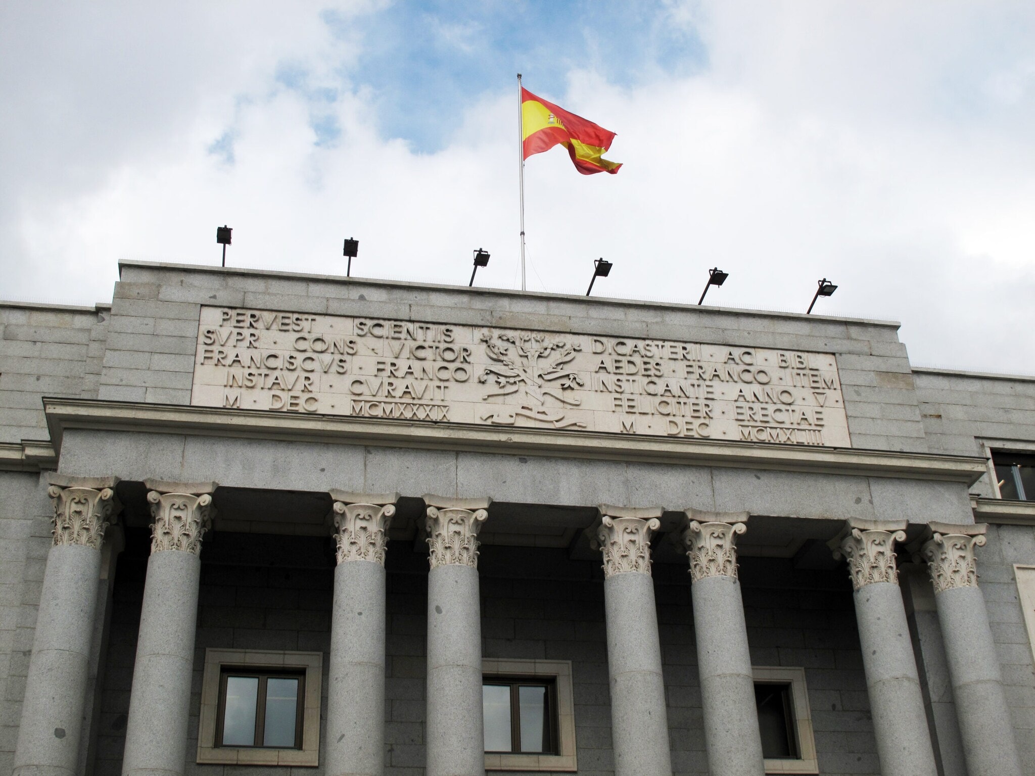 Mit Säulen geschmückter Eingangsbereich eines monumentalen Gebäudes mit Inschrift und wehender spanischer Flagge auf dem Dach Mit Säulen geschmückter Eingangsbereich eines monumentalen Gebäudes mit Inschrift und wehender spanischer Flagge auf dem Dach