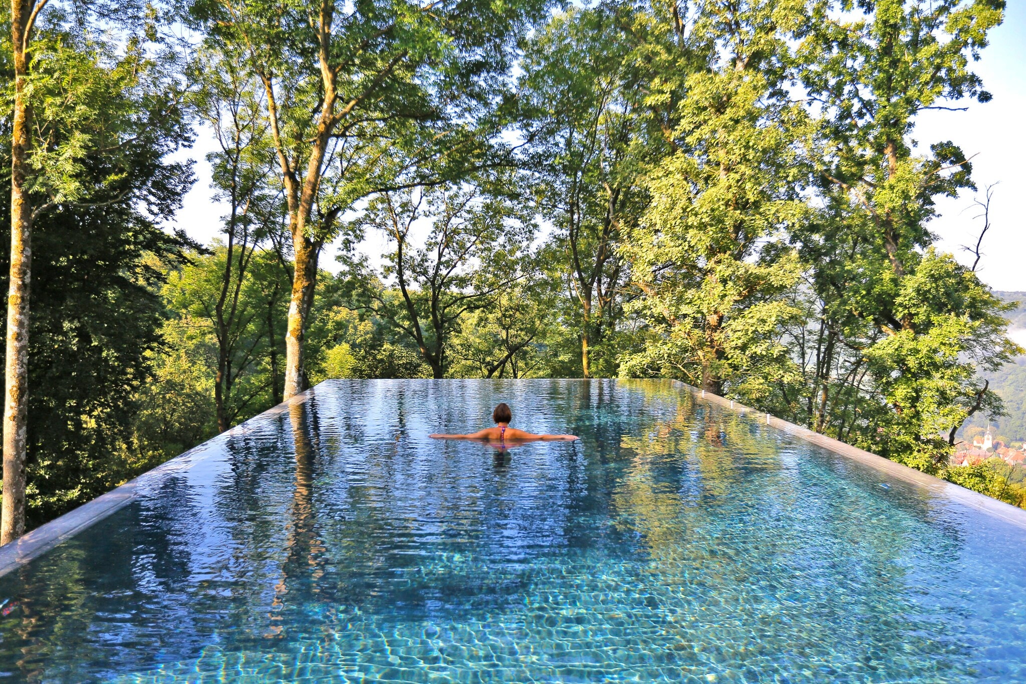 Rückansicht einer Frau im Infinity-Pool, im Hintergrund Baumkronen Rückansicht einer Frau im Infinity-Pool, im Hintergrund Baumkronen