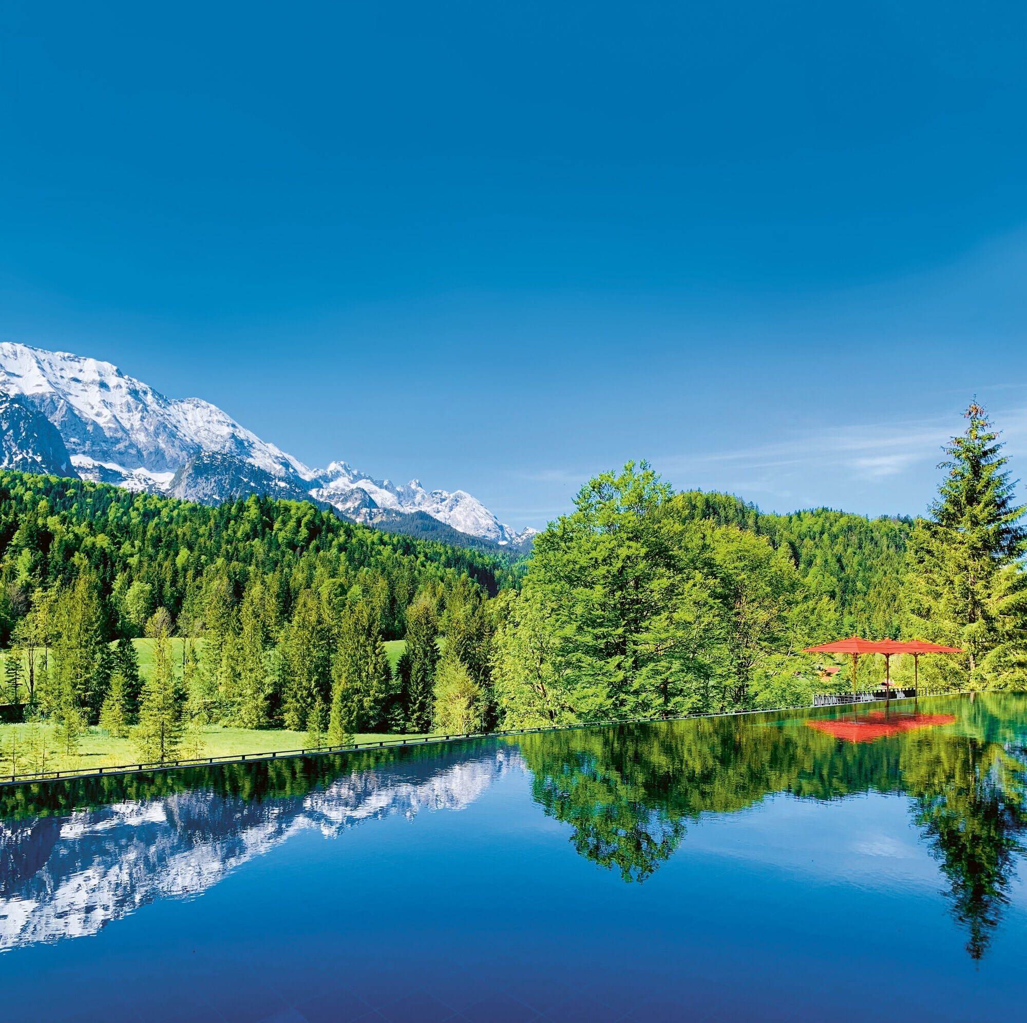 Infinitypool, in dem sich grüner Wald und Bergpanorama spiegeln, am Rande drei Sonnenliegen