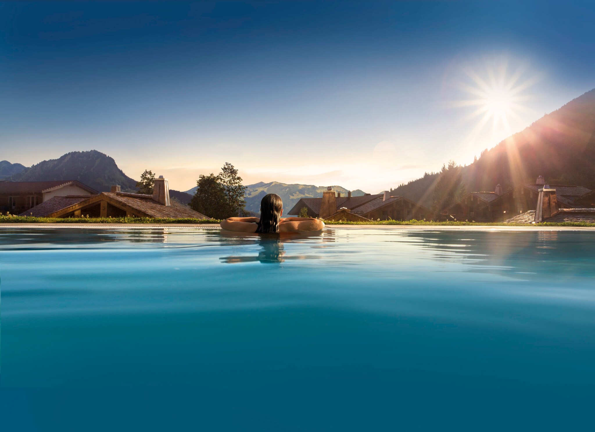 Rückansicht einer Frau im Infinitypool mit Blick über den Ort auf die Berge bei Sonnenschein