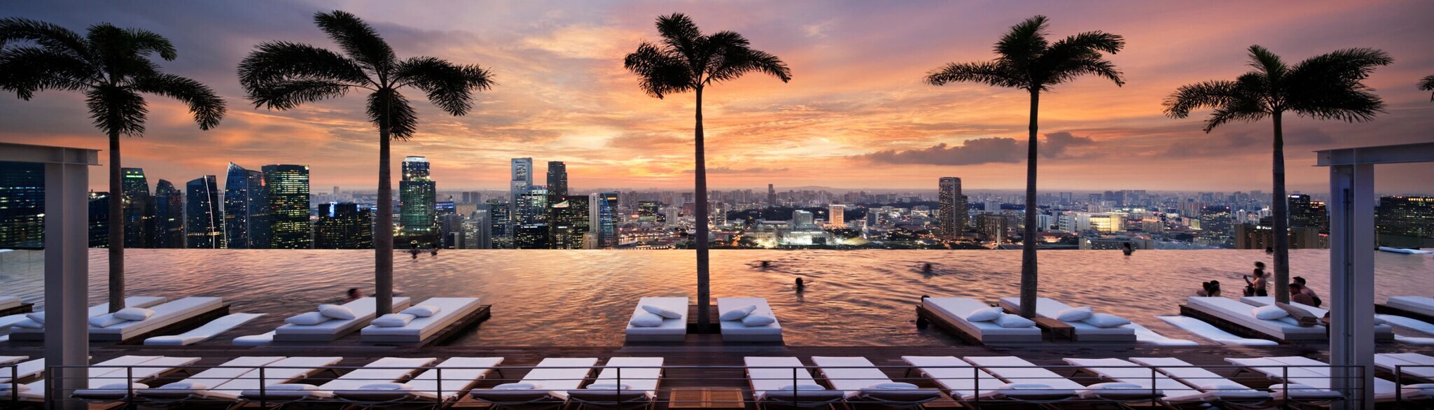 Ein endlos scheinender Pool mit Blick auf die Skyline Singapurs, mit Liegen und Palmen im Wasser