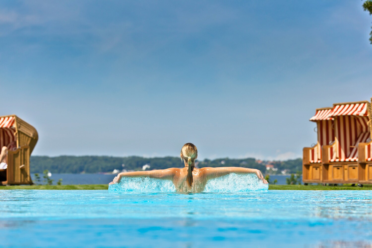 Rückansicht einer Frau im Pool mit ausgestreckten Armen, im Hintergrund Strandkörbe vor einer Meeresbucht