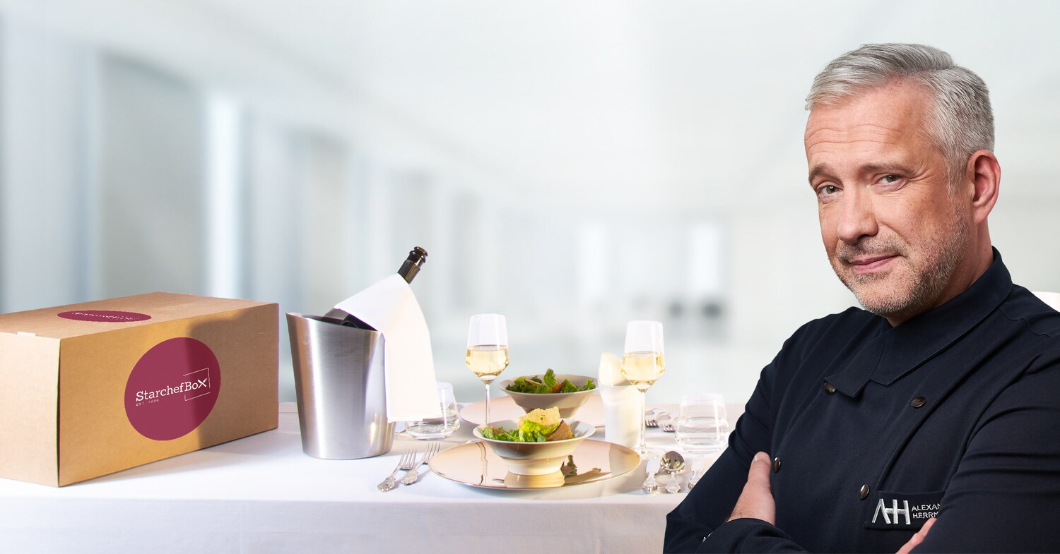 otomontage einer Karton-Box mit Logo auf einem elegant gedeckten Tisch neben einem Sektkühler in einem hellen Raum, im Vordergrund Sternekoch Alexander Herrmann mit Kochjacke, der in die Kamera blickt otomontage einer Karton-Box mit Logo auf einem elegant gedeckten Tisch neben einem Sektkühler in einem hellen Raum, im Vordergrund Sternekoch Alexander Herrmann mit Kochjacke, der in die Kamera blickt