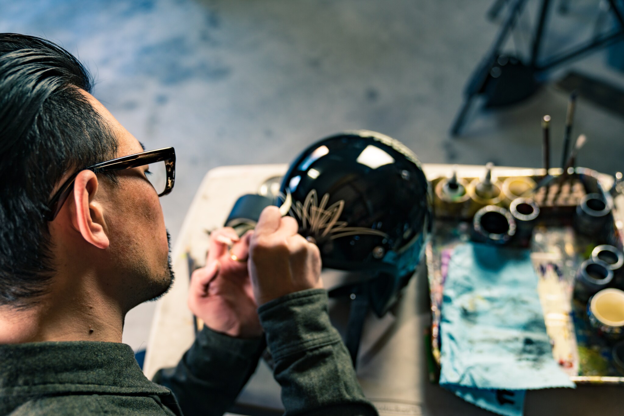 Blick über die Schulter eines Mannes, der einen schwarzen Motorradhelm mit goldenen Elementen bemalt, daneben Malutensilien in einem Atelier