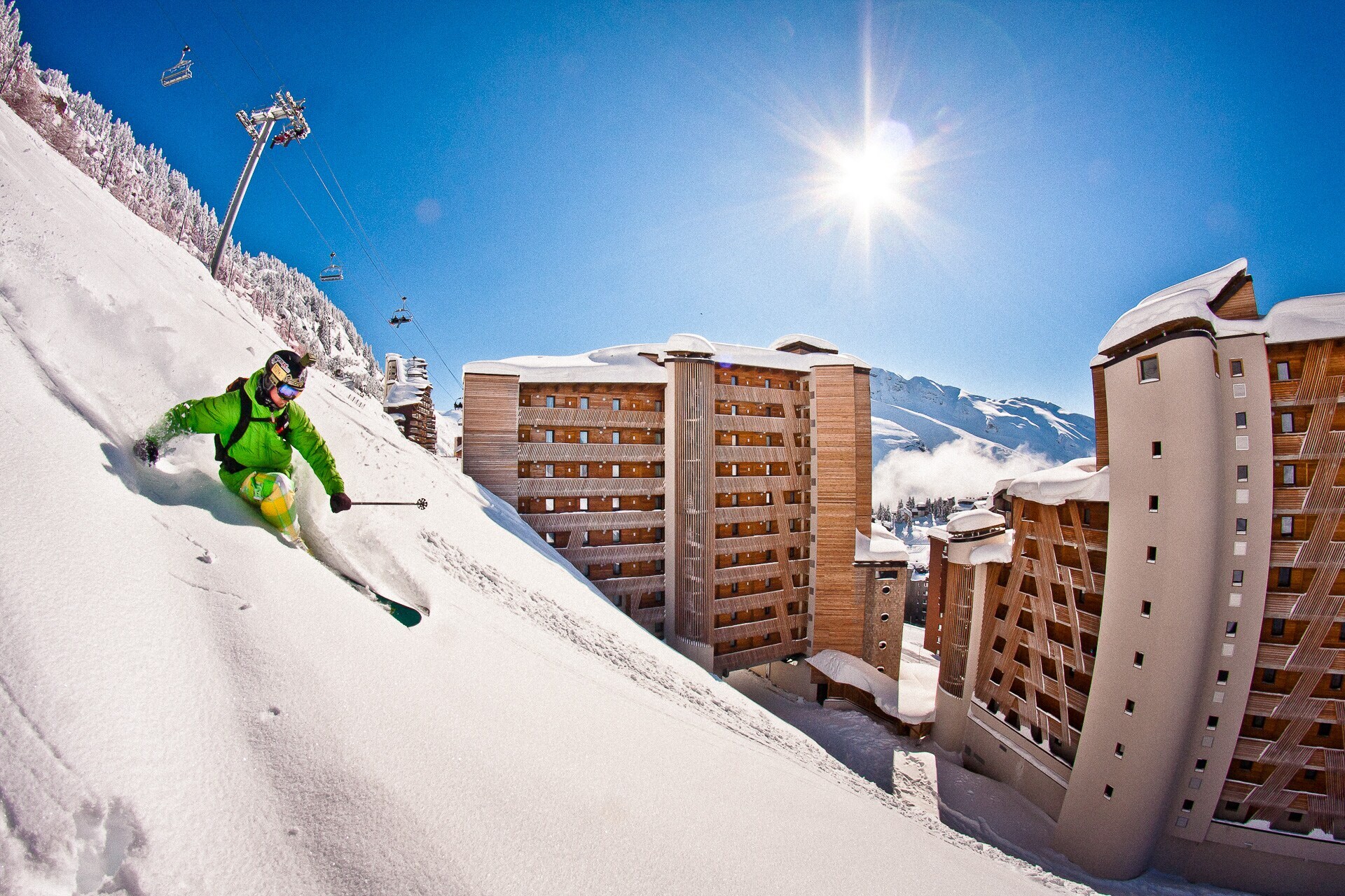 Eine Person mit Ski fährt einen steilen Hang hinab, im Hintergrund sind Häuser und Berge.