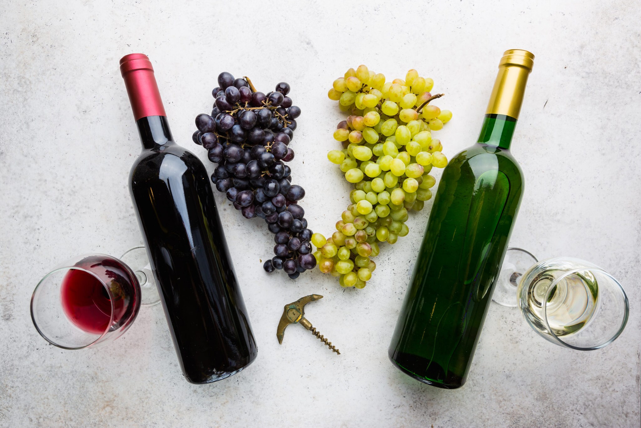 Je eine Flasche und ein Glas Rotwein und Weißwein auf einer hellen Fläche, dazwischen liegen Trauben. Je eine Flasche und ein Glas Rotwein und Weißwein auf einer hellen Fläche, dazwischen liegen Trauben.