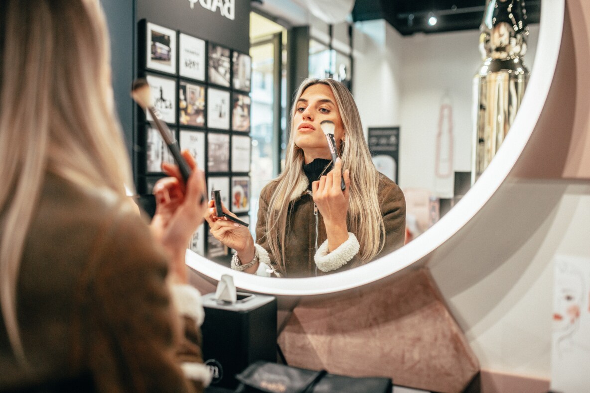 Eine Frau steht vor einem großen runden Spiegel und pudert sich das Gesicht