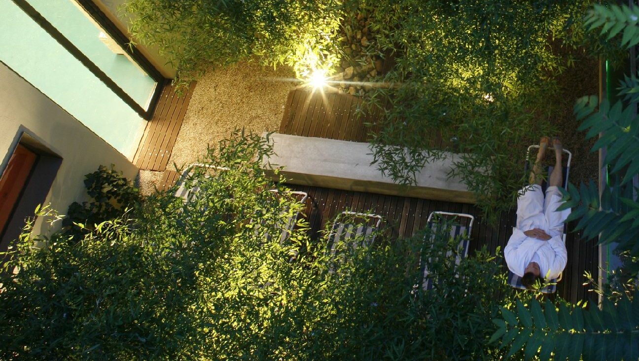 Eine Person liegt im Bademantel auf einer Liege in einem Patio mit grünen Pflanzen aus der Vogelperspektive Eine Person liegt im Bademantel auf einer Liege in einem Patio mit grünen Pflanzen aus der Vogelperspektive