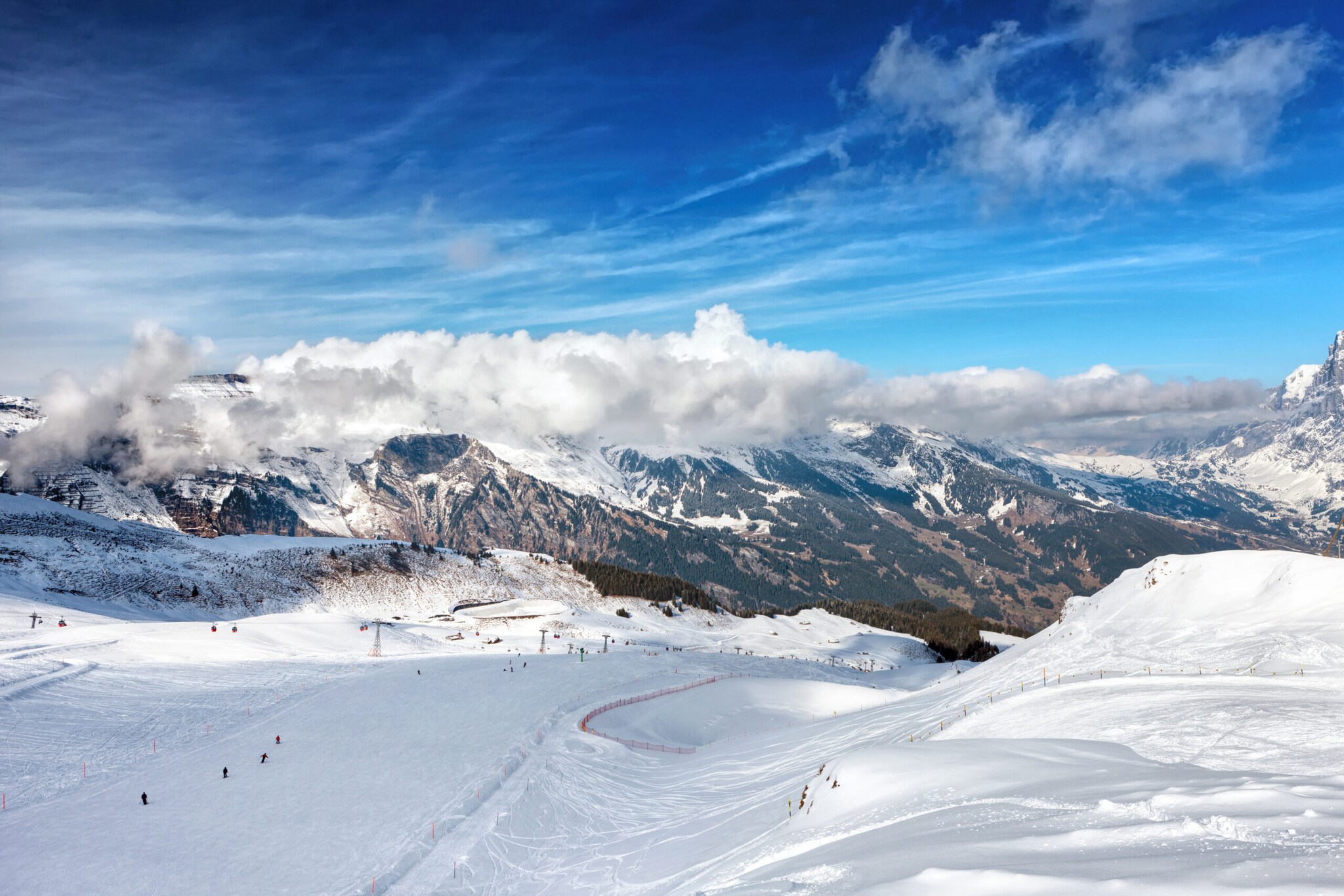 Blick von oben auf eine Piste mit Skifahrer:innen, im Hintergrund Berge