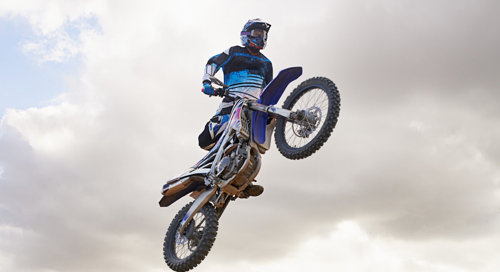 Ein Motocrossfahrer fliegt durch die Luft, im Hintergrund ein wolkenbehangener Himmel und ein Wald Ein Motocrossfahrer fliegt durch die Luft, im Hintergrund ein wolkenbehangener Himmel