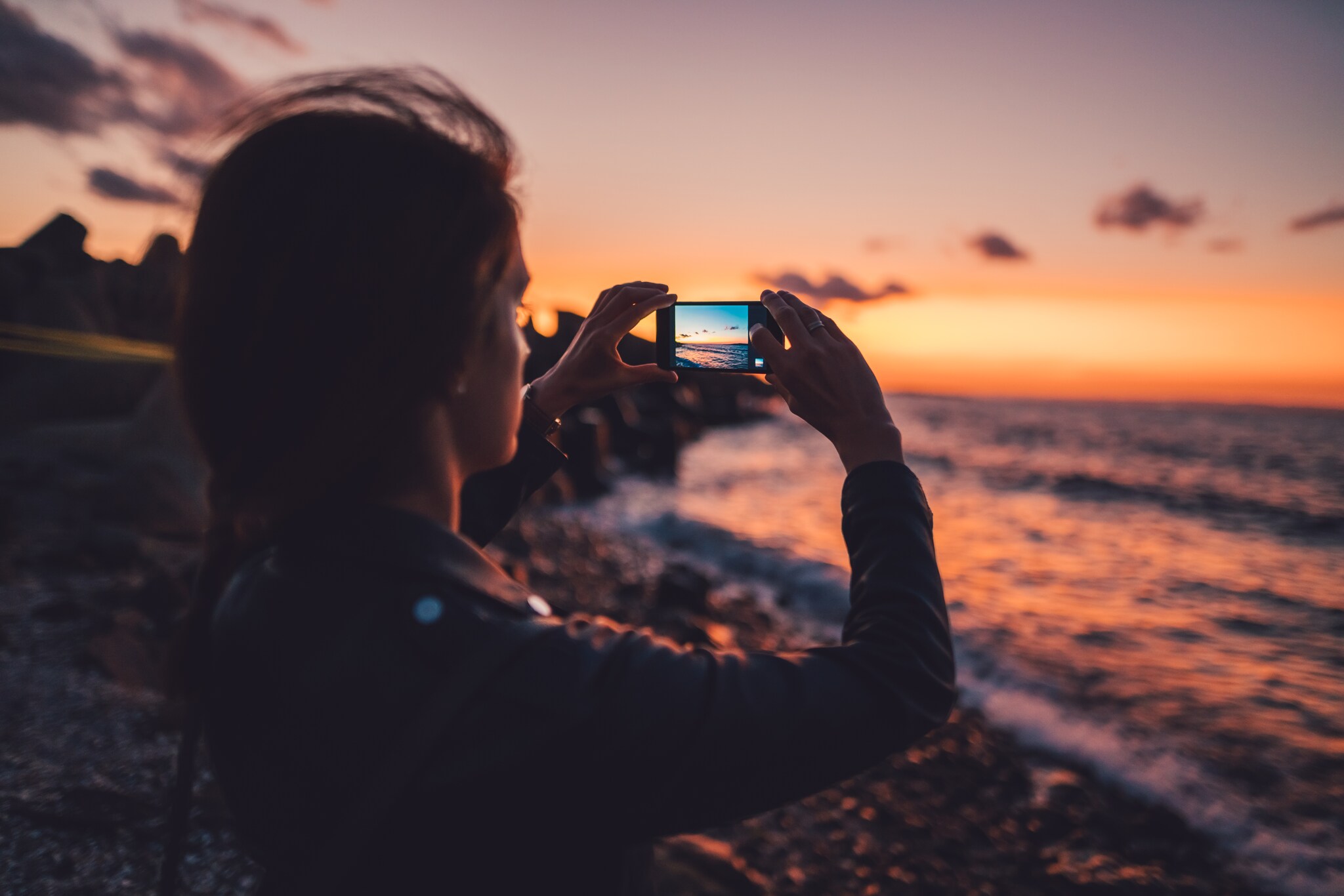 Eine junge Frau fotografiert einen Sonnenuntergang am Meer mit dem Handy