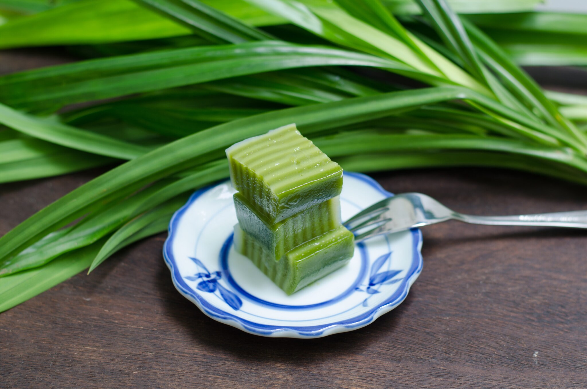 Ein kleiner Teller mit einem grünen Dessert, daneben frische Pandanblätter